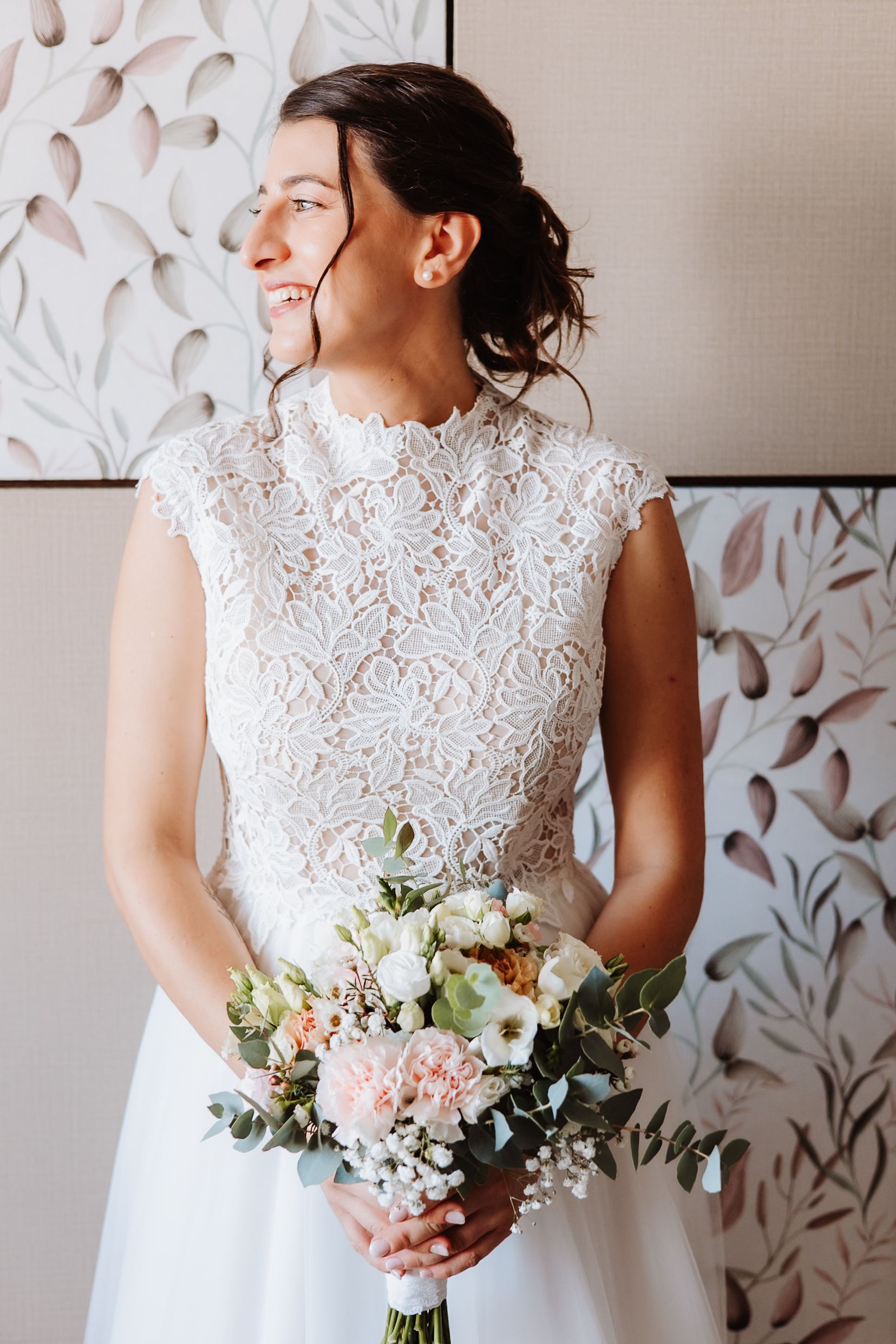 Bride in elegant lace cap sleeve wedding dress holds delicate bouquet of white and pink flowers against textured wall.