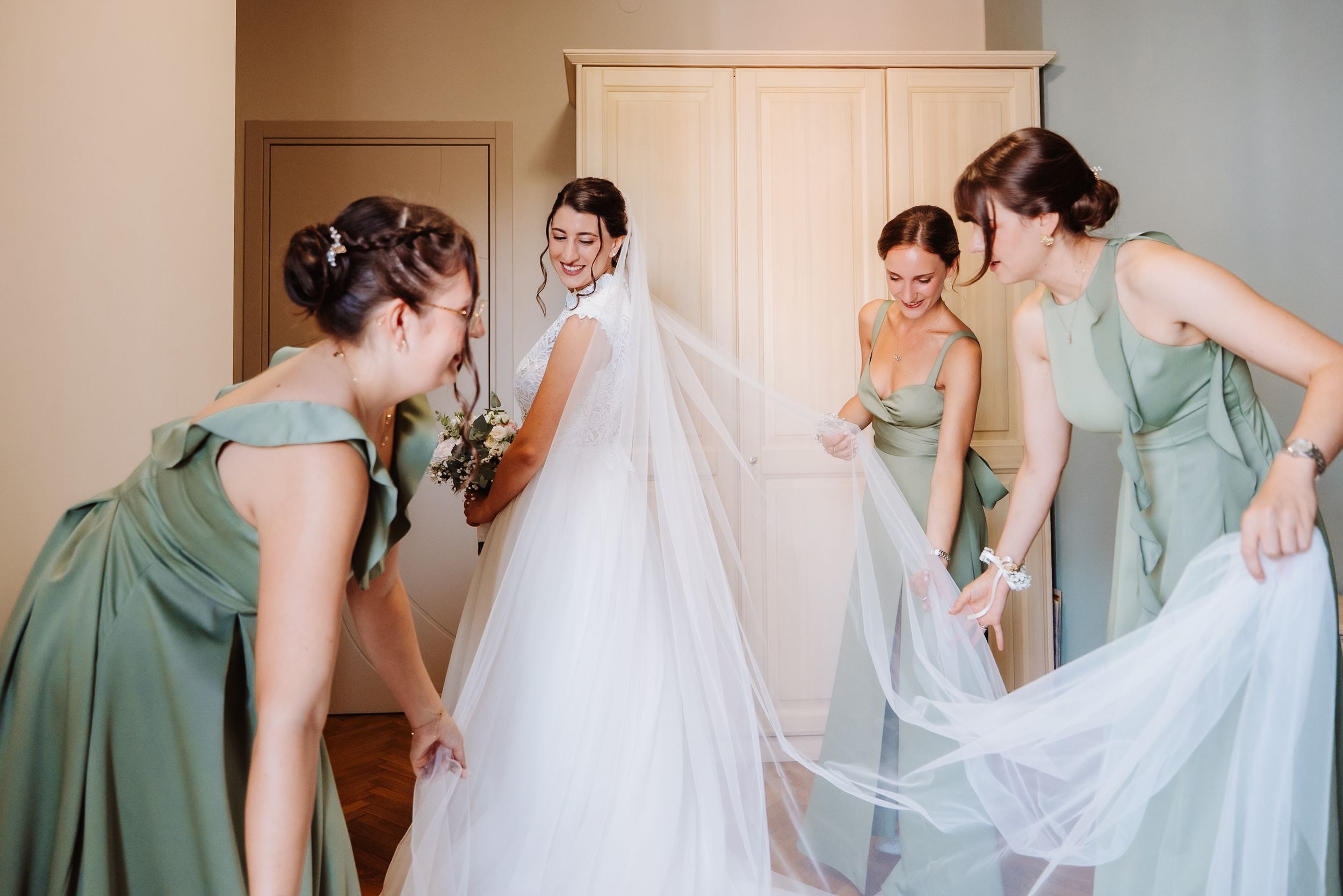 Bridesmaids in sage green dresses help the bride get ready for her wedding day in a sunlit room.