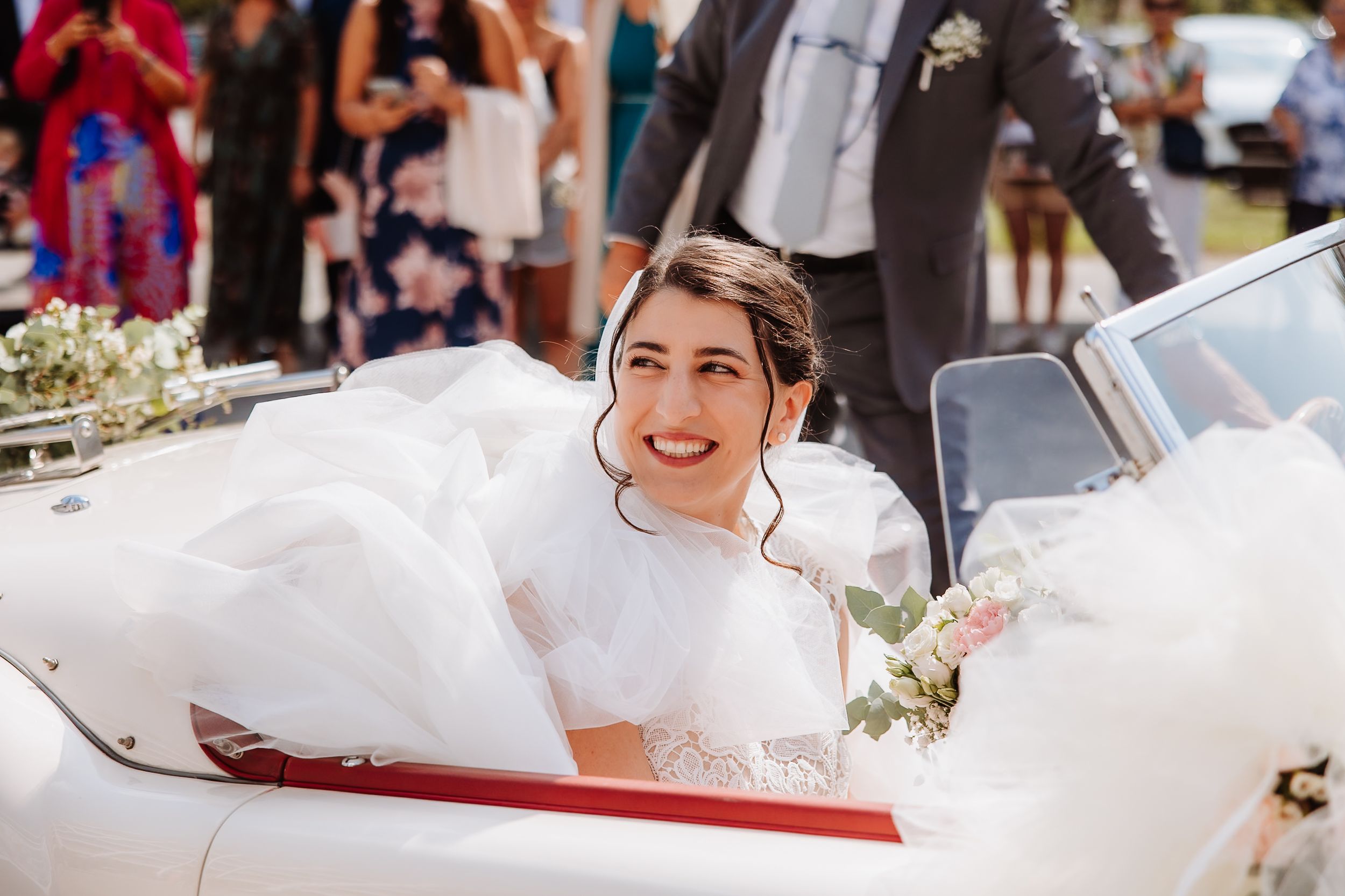 A bride in a white wedding gown sits in a classic red convertible car during a sunny outdoor celebration.