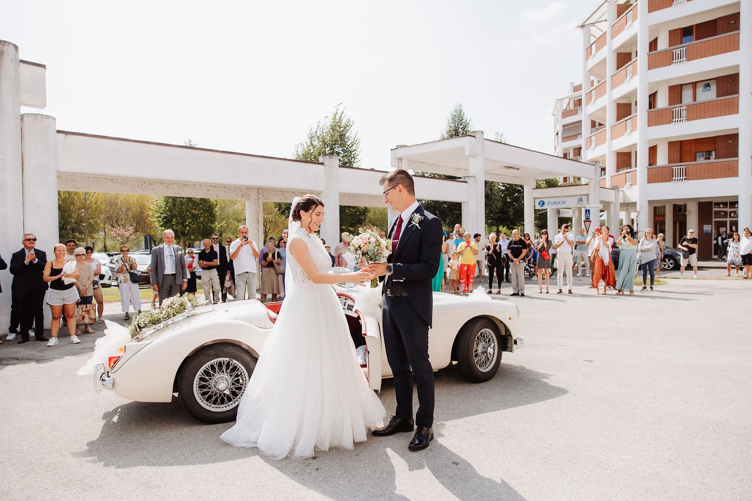 A classic white vintage car parked outside during a wedding celebration with guests gathered around.