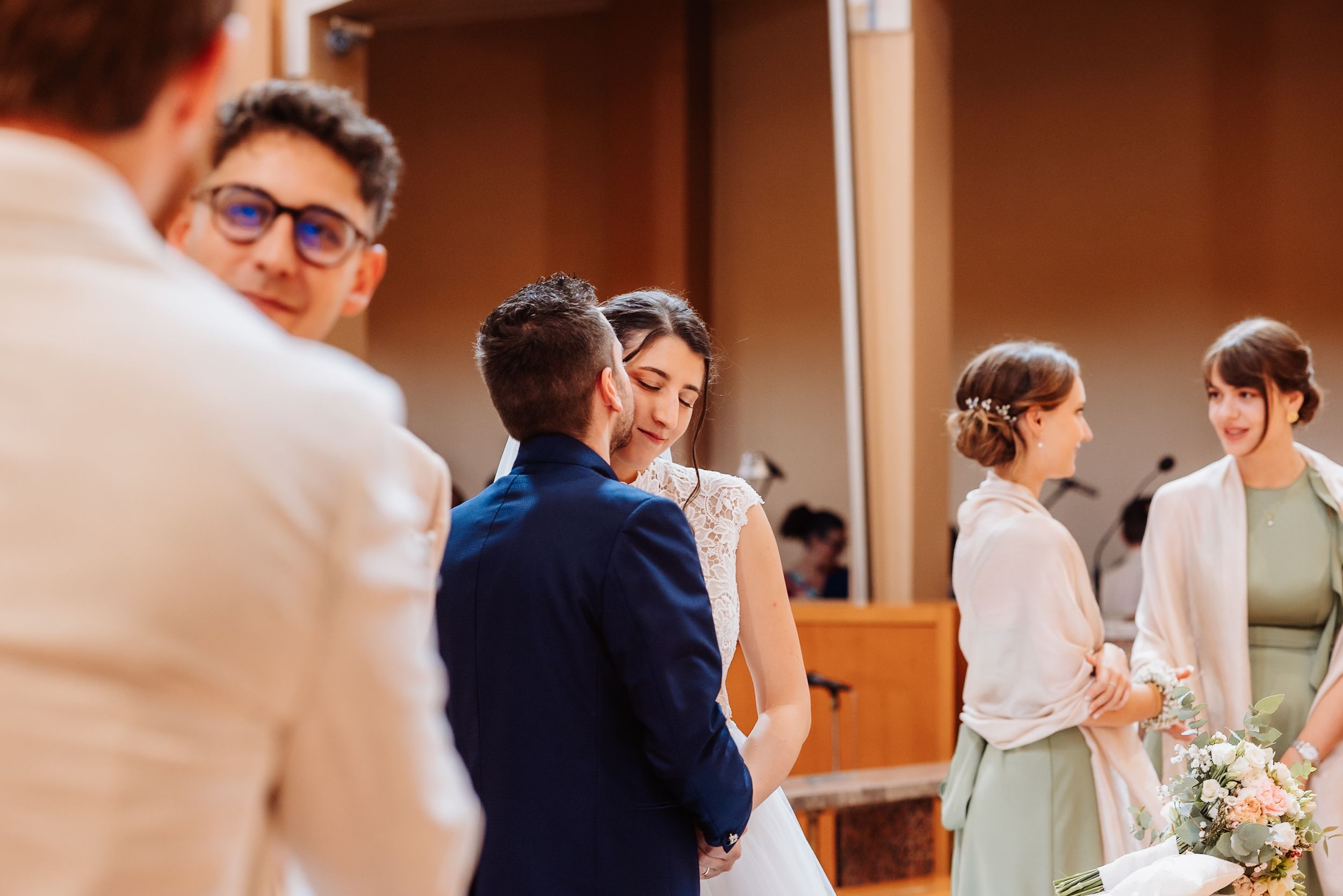 Wedding ceremony attendees gathered in a warm-lit indoor venue with soft peach tones.