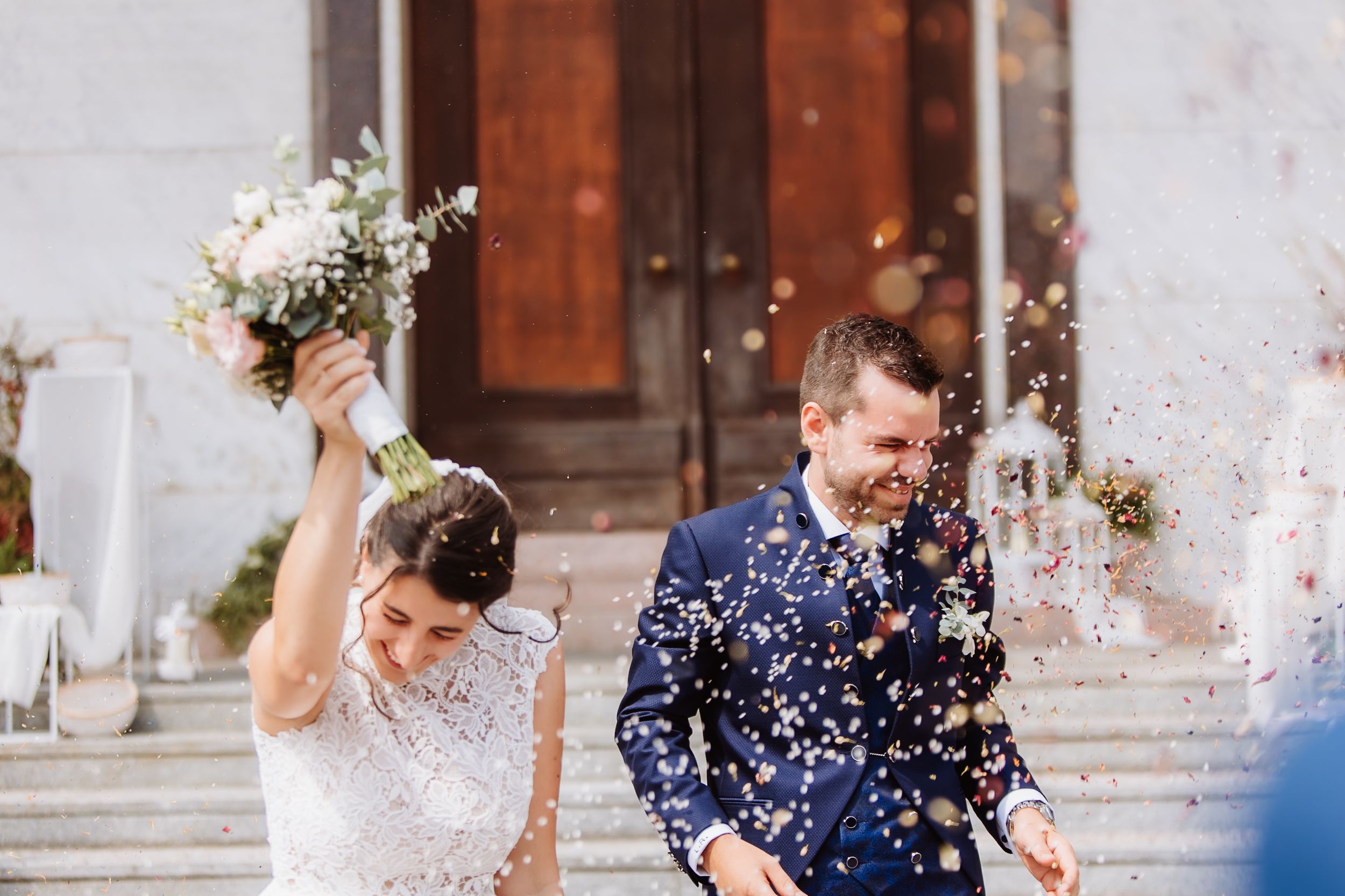 A joyful couple exits their wedding ceremony while guests shower them with white confetti on church steps.