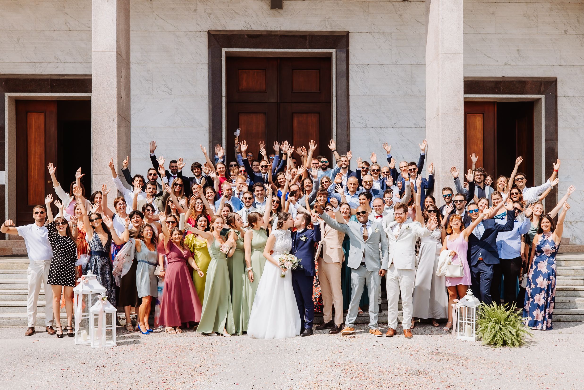 Large group of wedding guests pose together for a panoramic photo outside a white stone building with wooden doors.