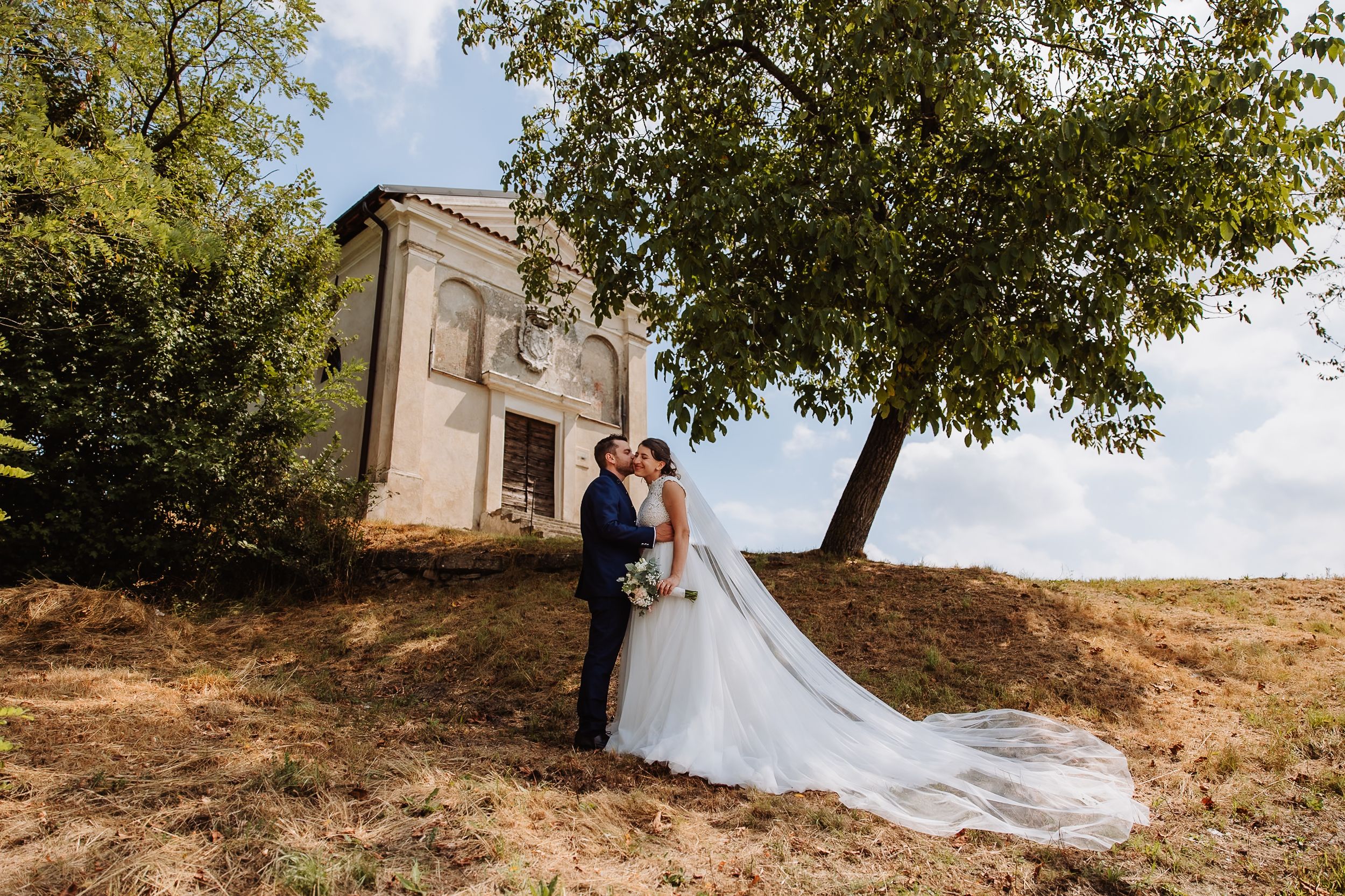 Romantic wedding couple embraces on a rustic hillside with trees and natural scenery in warm sunlight.