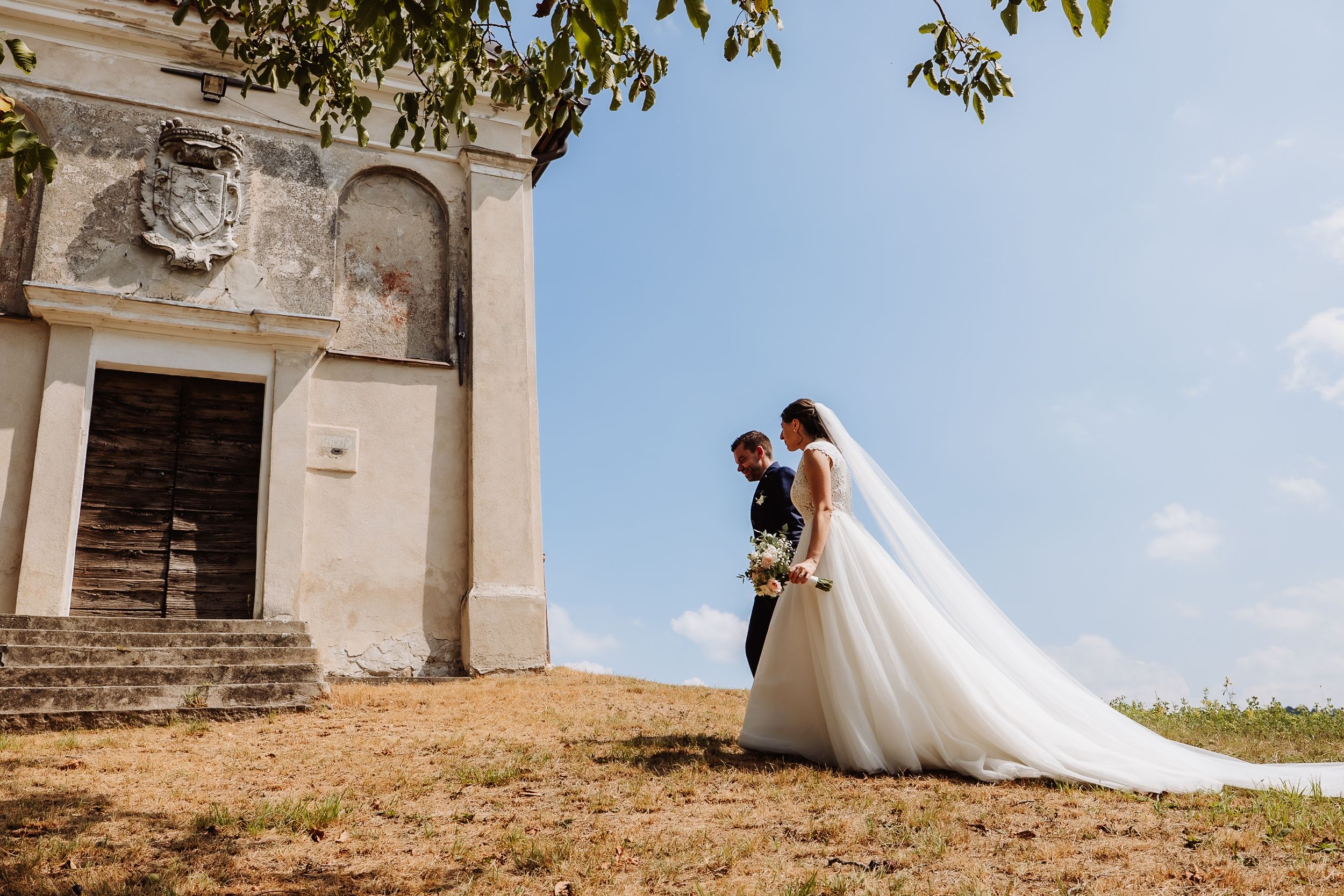 A couple in wedding attire embrace outside a historic stone church on a sunny hilltop.