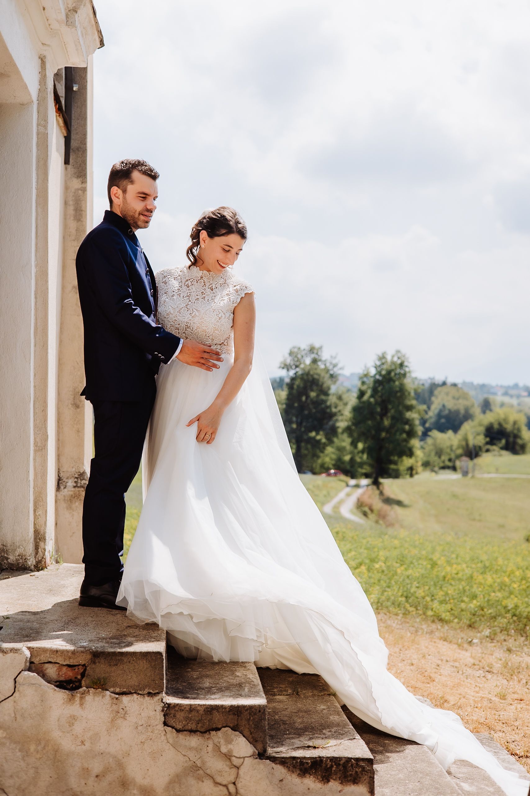 Romantic wedding couple poses on rustic stone steps with flowing white dress against countryside backdrop.