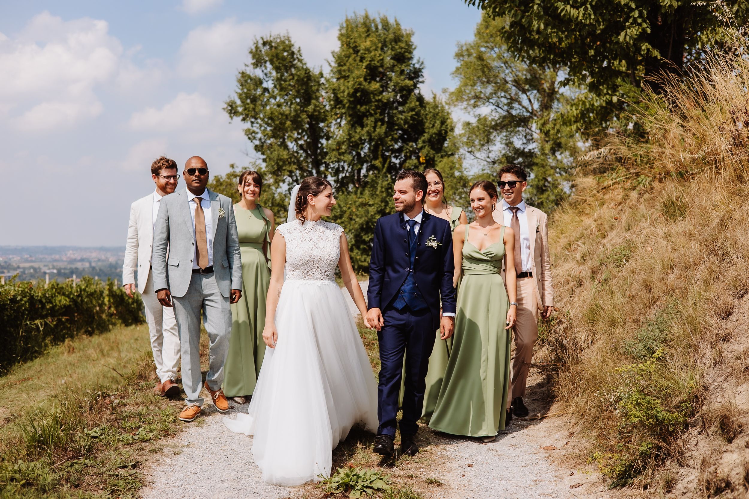 Wedding party walks along scenic hillside path on sunny summer day with trees and blue sky in background.