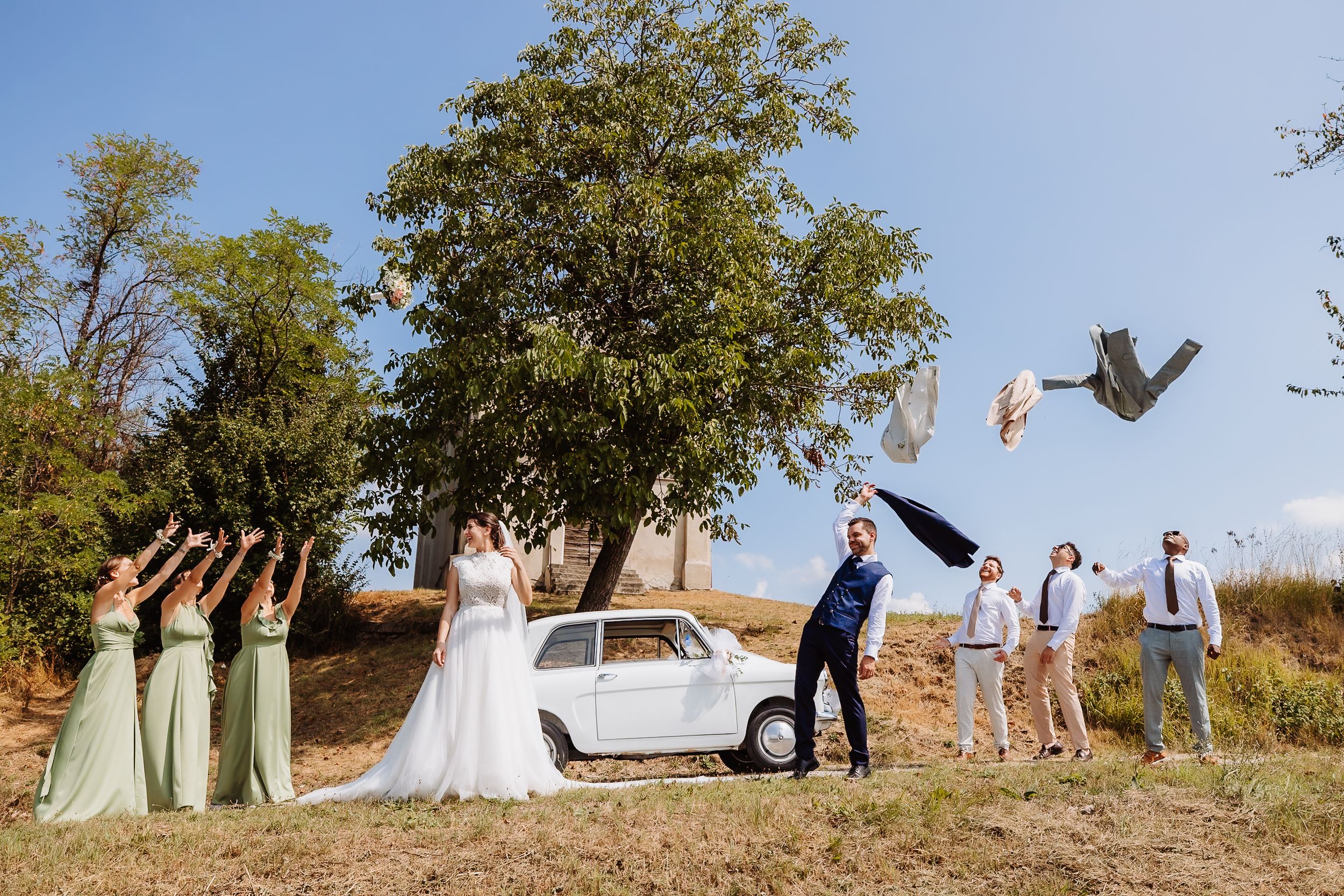 Wedding party in sage green and white dresses pose playfully on a grassy hillside during sunset.