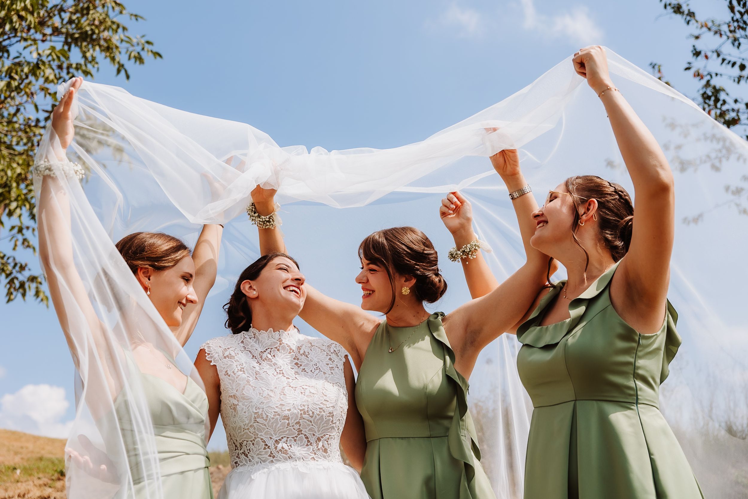 A group in sage green dresses and white gown celebrate with raised arms against a bright blue sky.