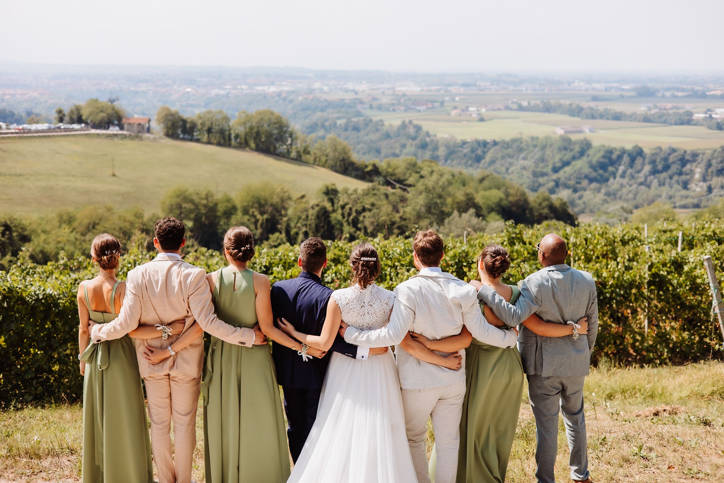 Wedding party stands arm in arm looking out over rolling green hills and scenic countryside landscape.