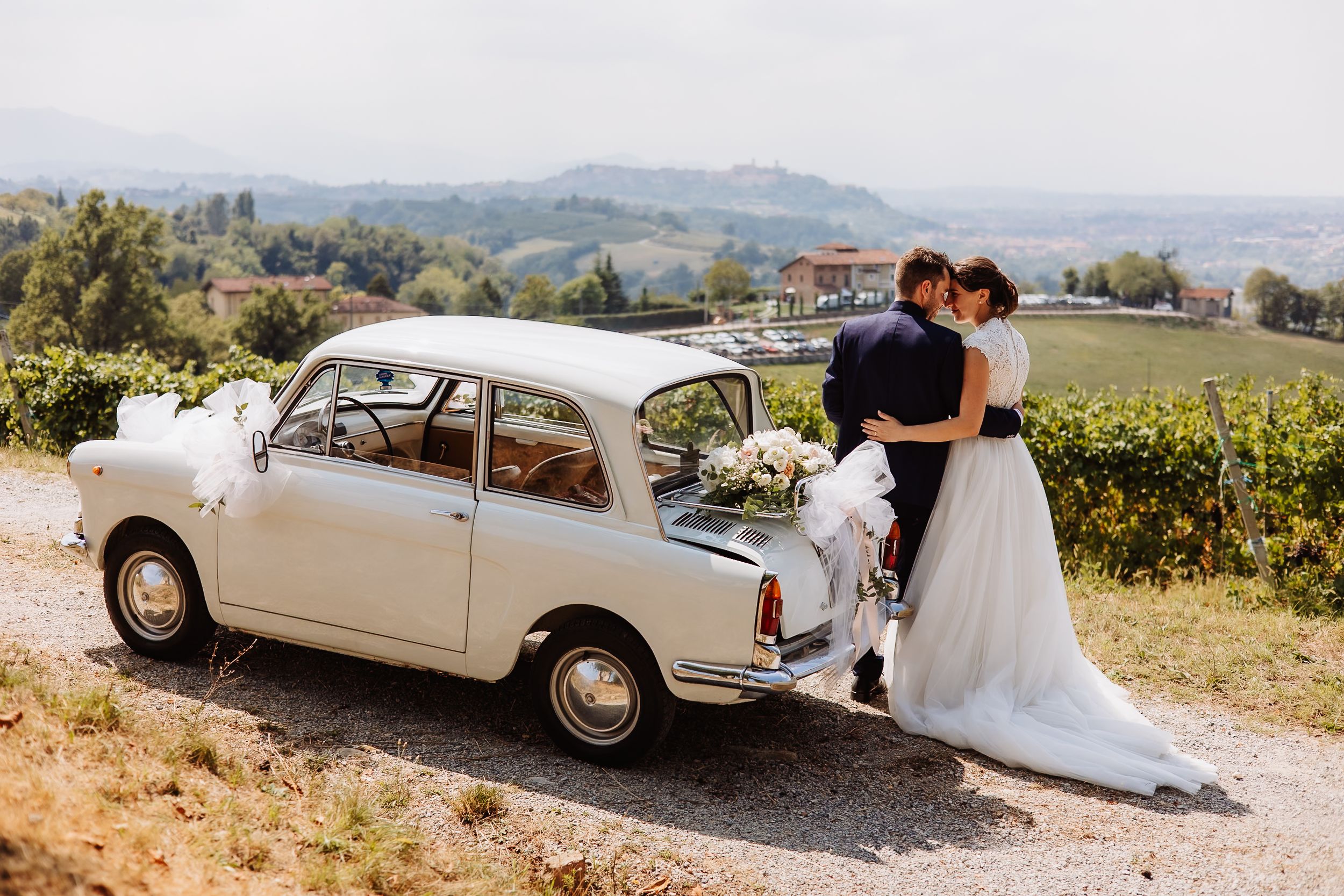 A vintage white Fiat 500 car parked on a hilltop with rolling vineyards in the scenic countryside.