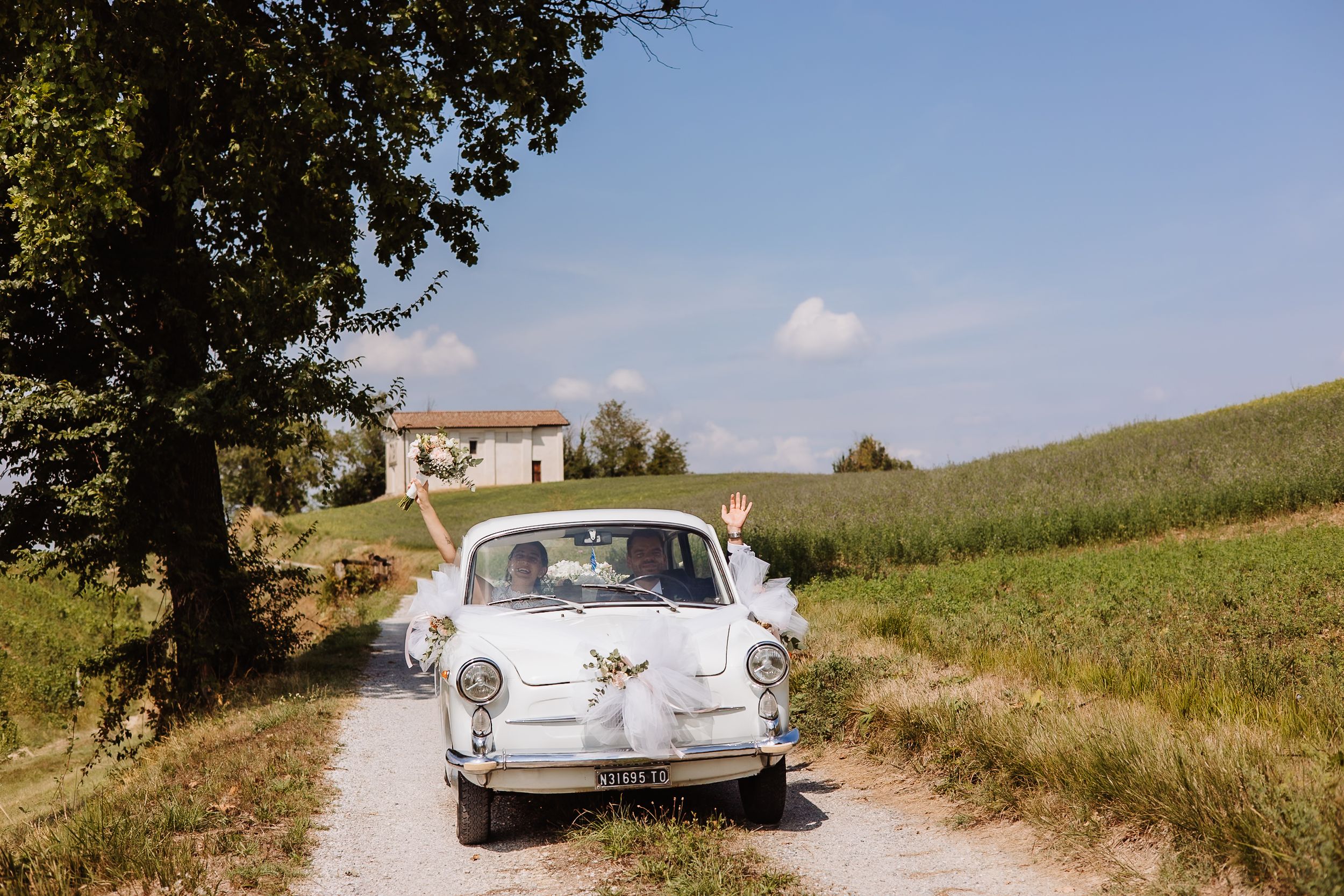A vintage white car travels down a scenic rural road through rolling countryside fields on a sunny day.