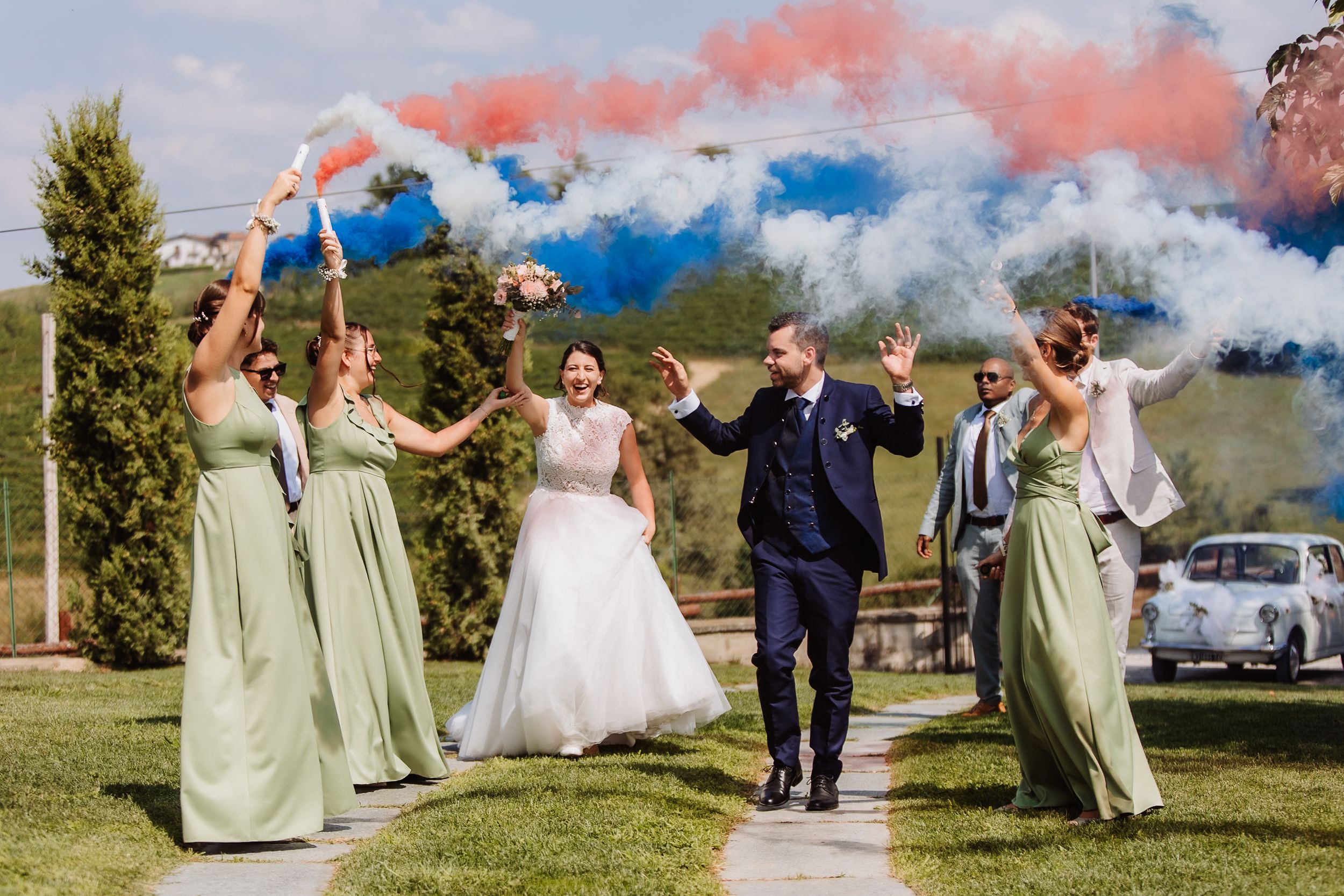 Wedding party celebrating with colorful smoke bombs creating red and blue clouds in an outdoor garden setting.