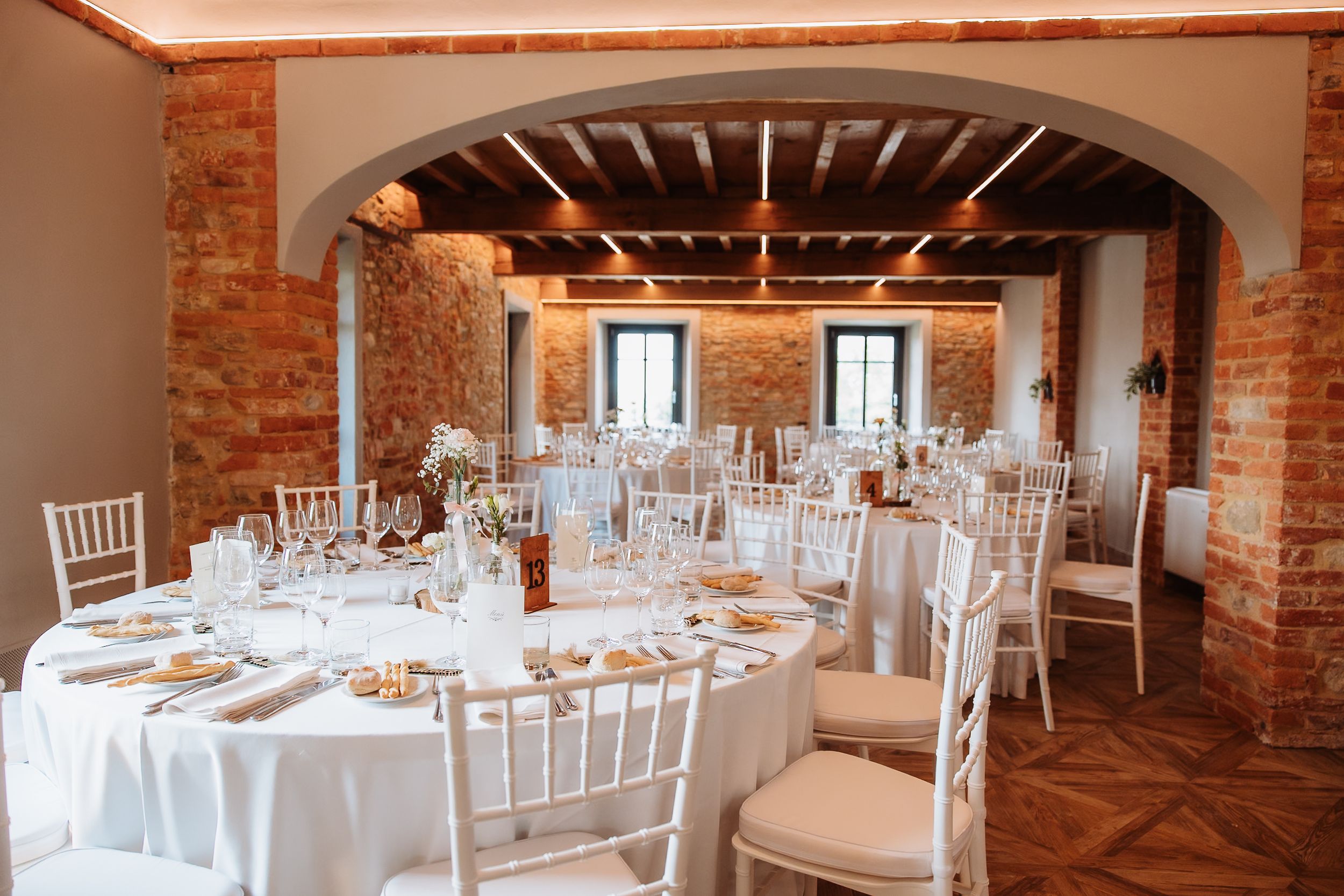 Elegant wedding reception venue with exposed brick walls, white chairs, and round tables set for dining.