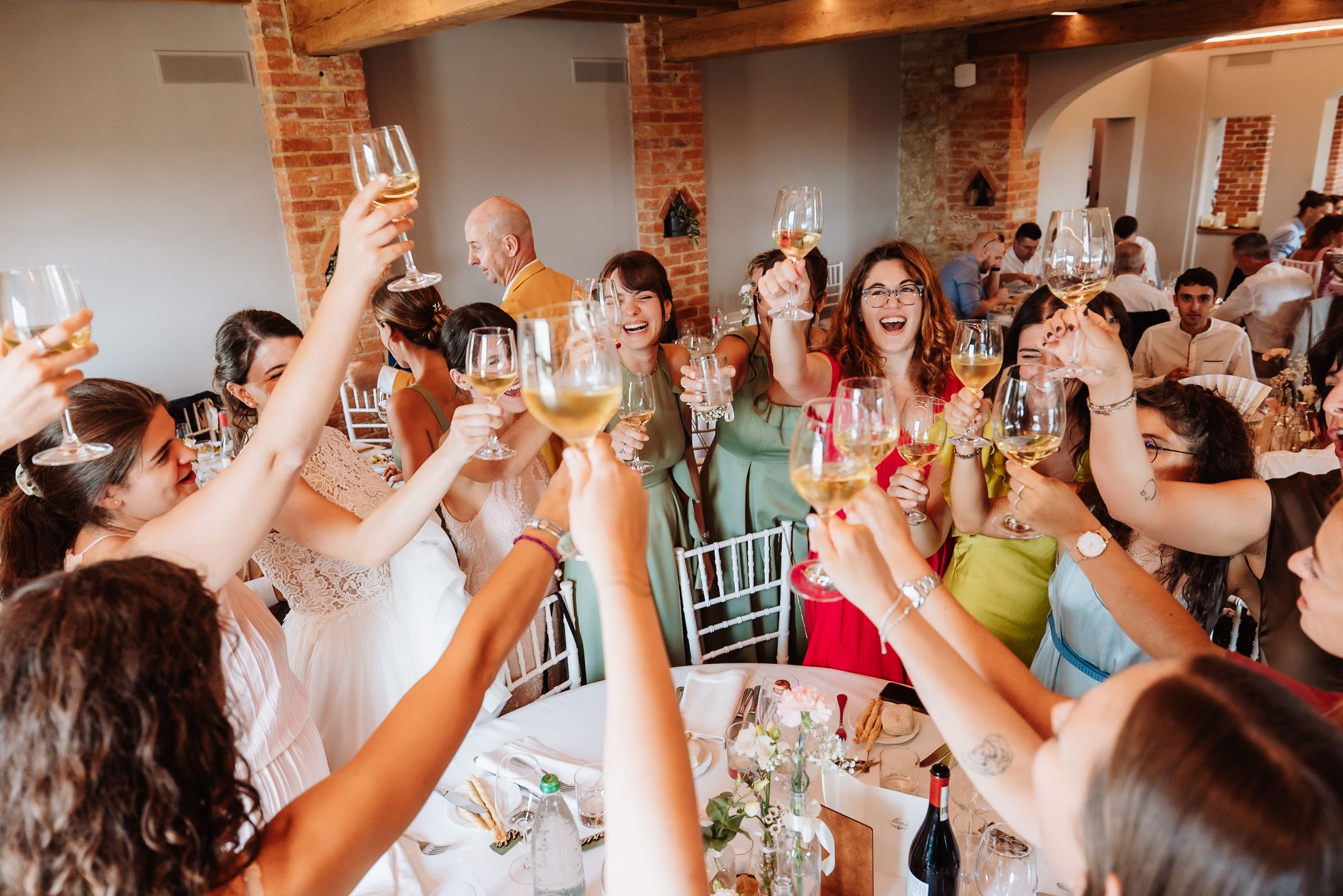 Wedding guests raise their glasses in celebration during a toast at a rustic brick-walled reception venue.