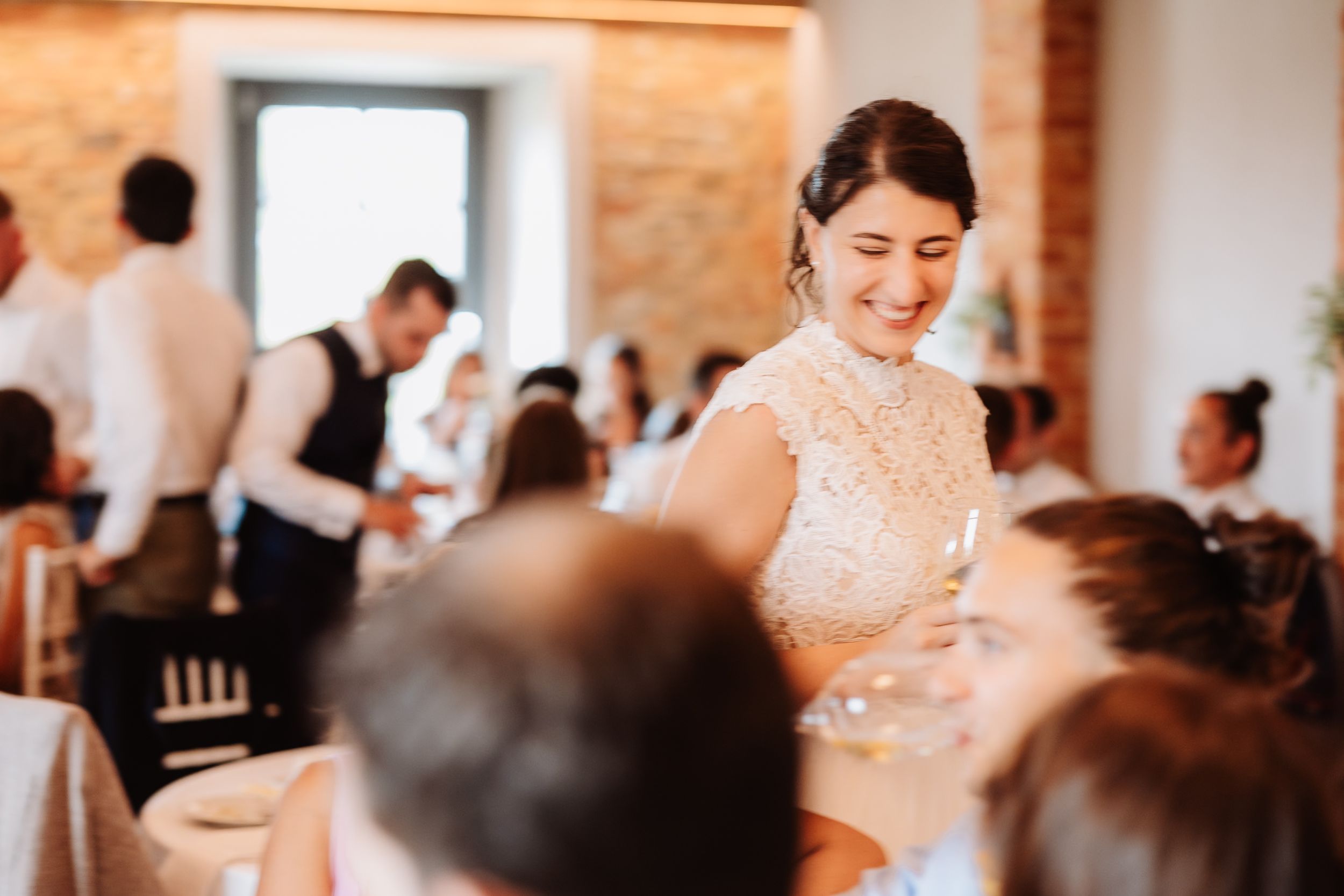 Wedding reception guests seated at round tables in a bright room with exposed brick walls and large windows.
