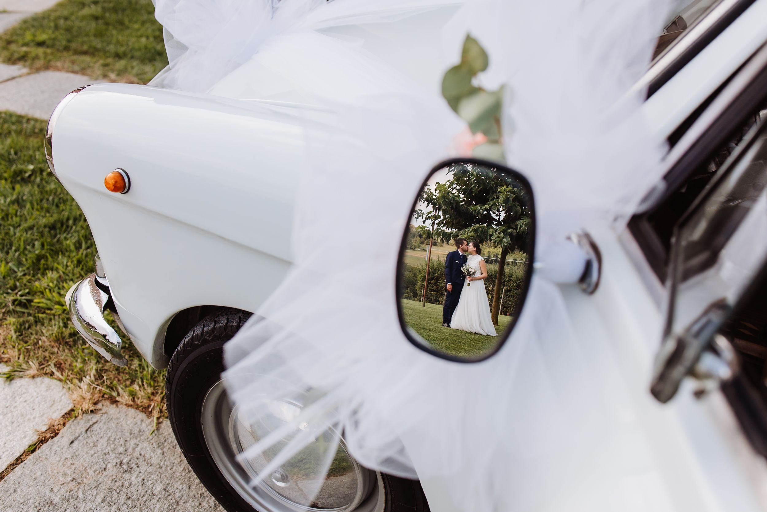 A close-up view of a white car's side mirror reflecting a sunny outdoor scene with trees and grass.