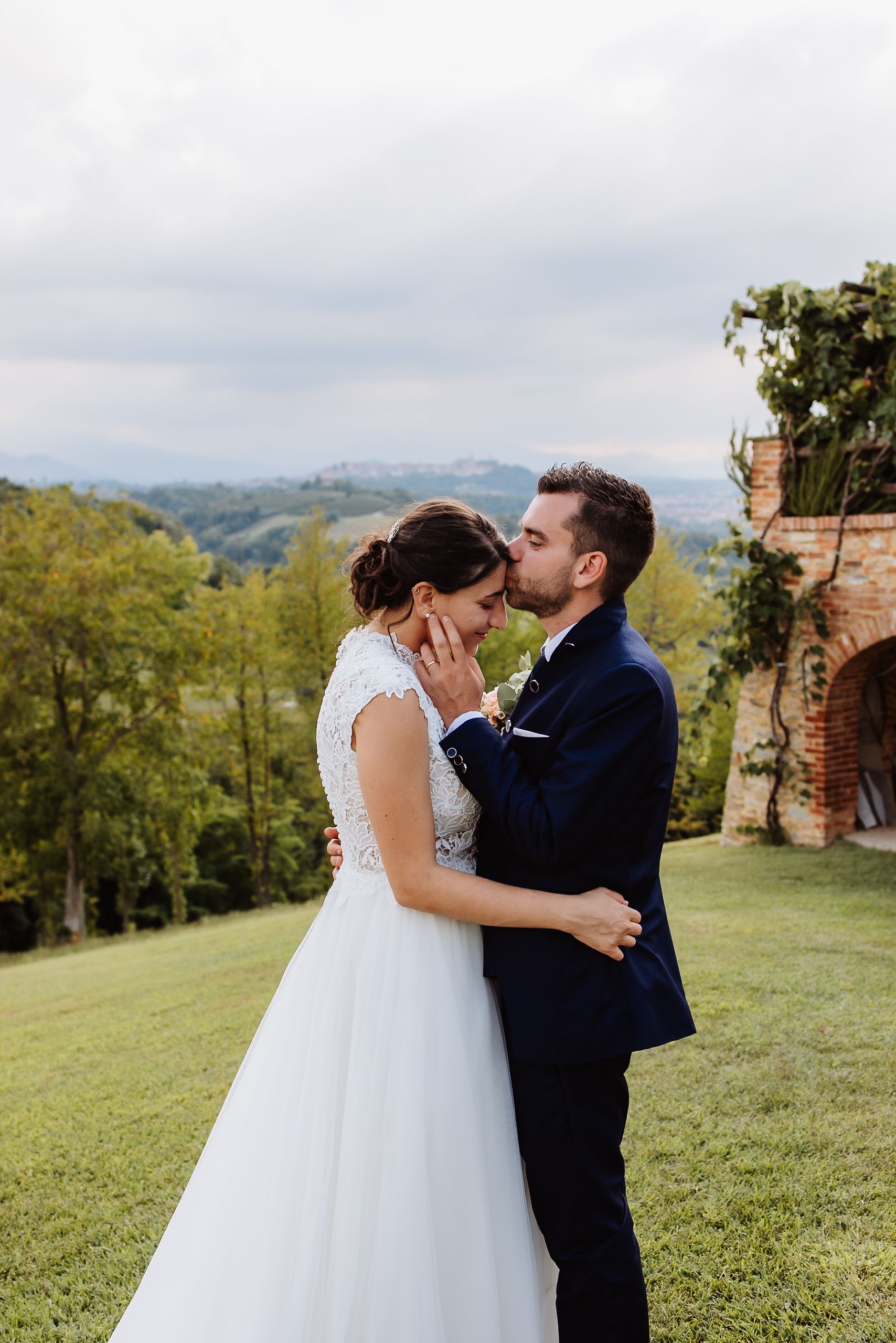 A couple shares a romantic kiss on a hilltop with rolling green countryside and trees in the background.