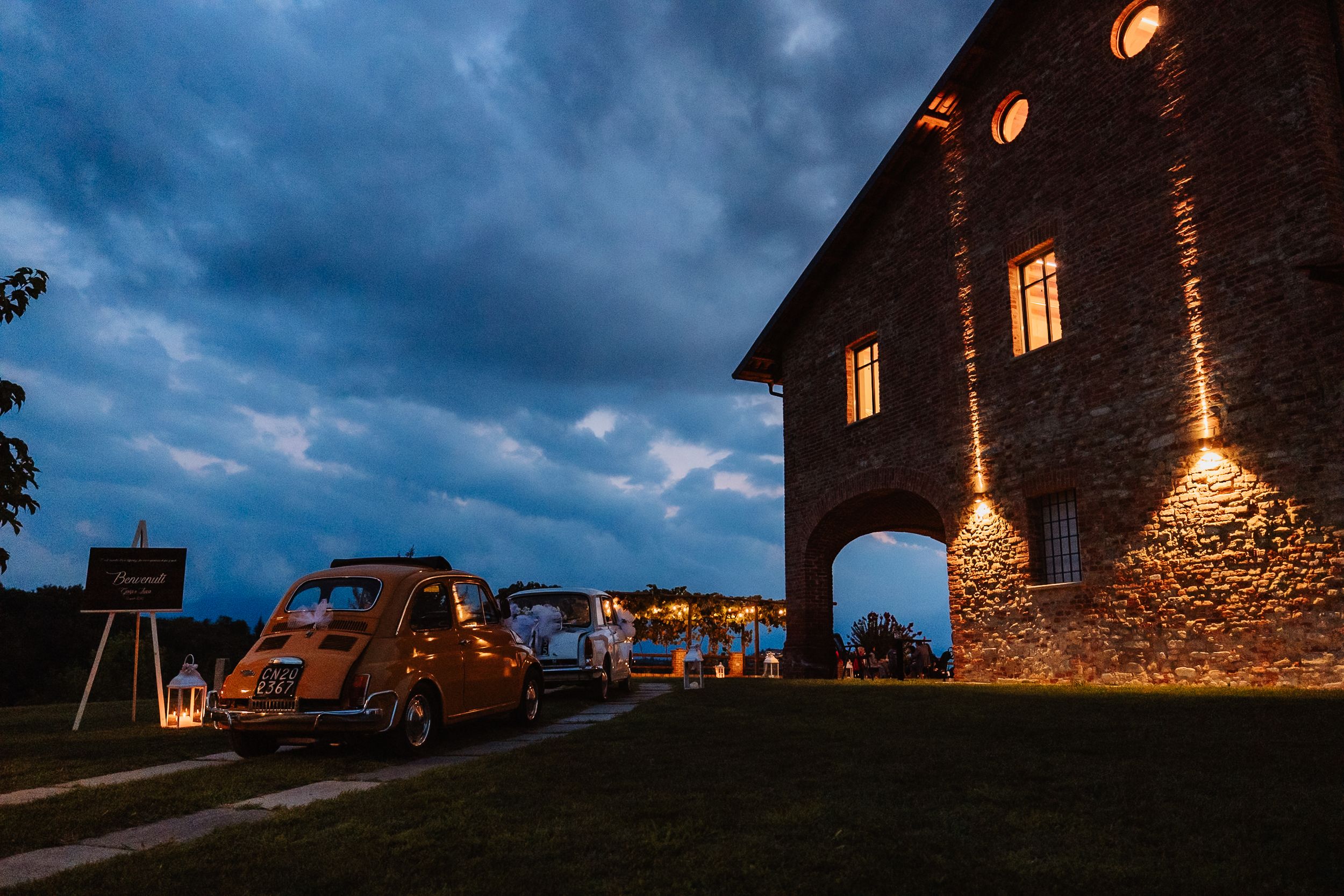 Panoramic dusk view of illuminated stone buildings and parked cars against a dramatic stormy blue sky.