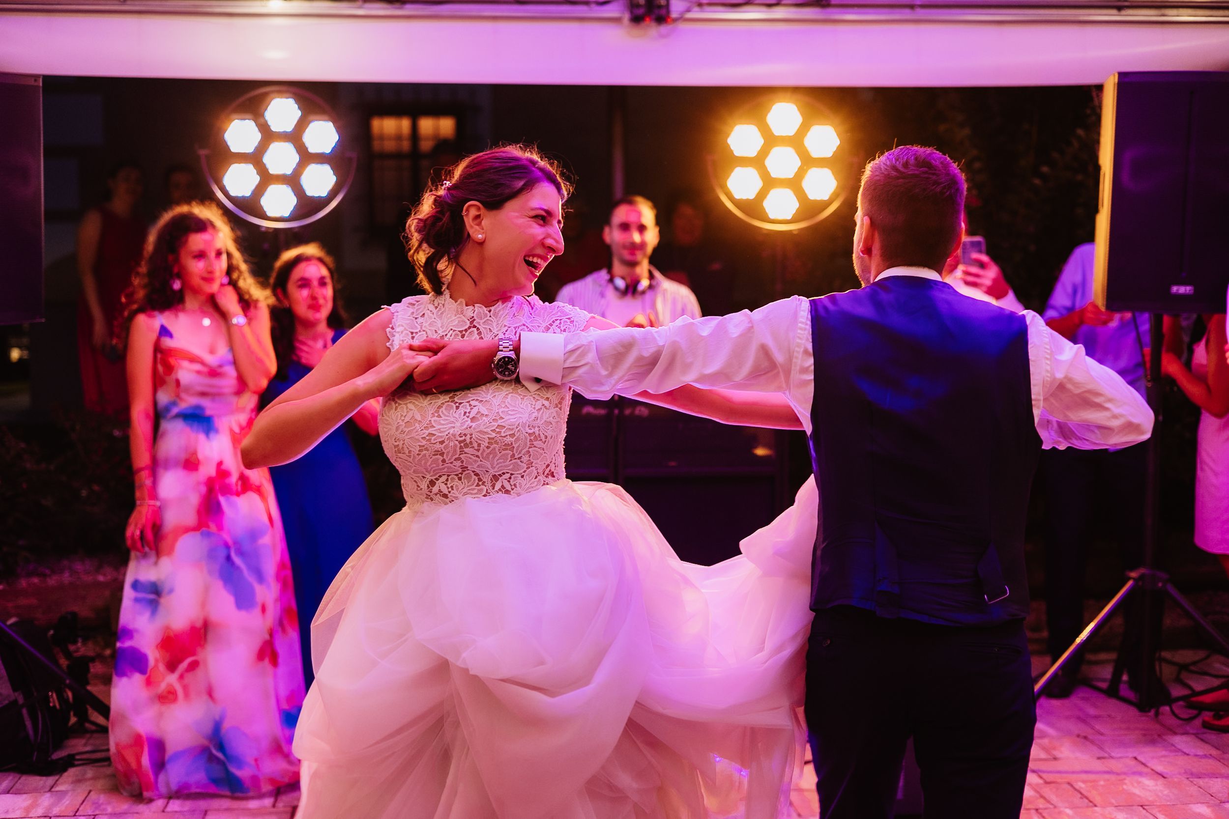 Wedding guests dance and celebrate on a wooden dance floor illuminated by warm stage lights at an evening reception.