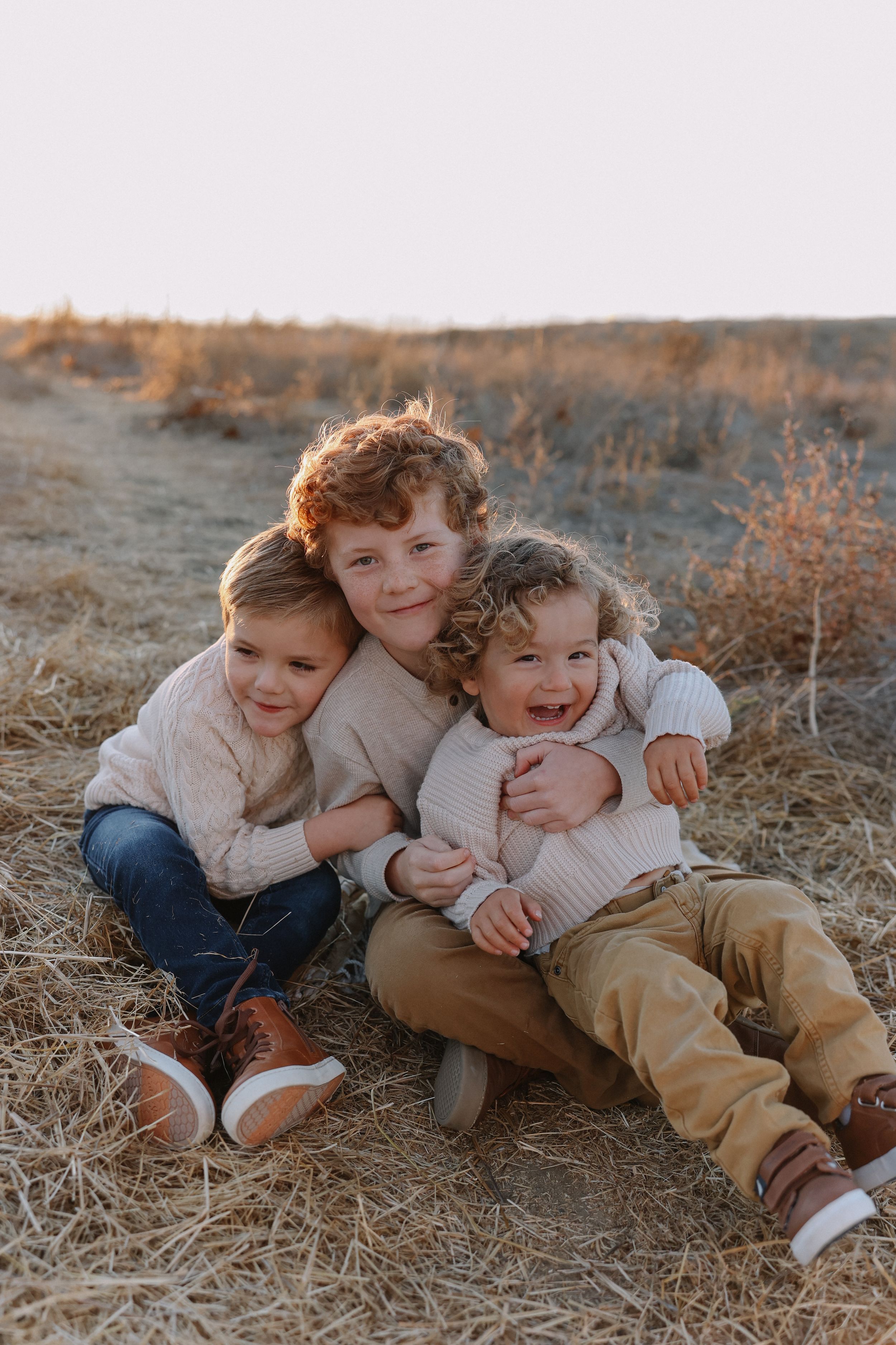 Three siblings in cream colored sweaters playfully cuddle together on dried grass during golden hour for family session