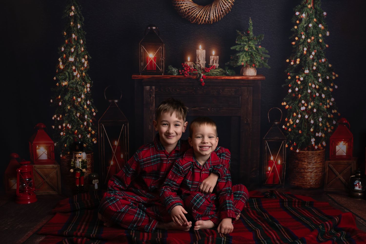 Children in red plaid shirts pose by decorated Christmas trees in a cozy holiday photo studio setting.