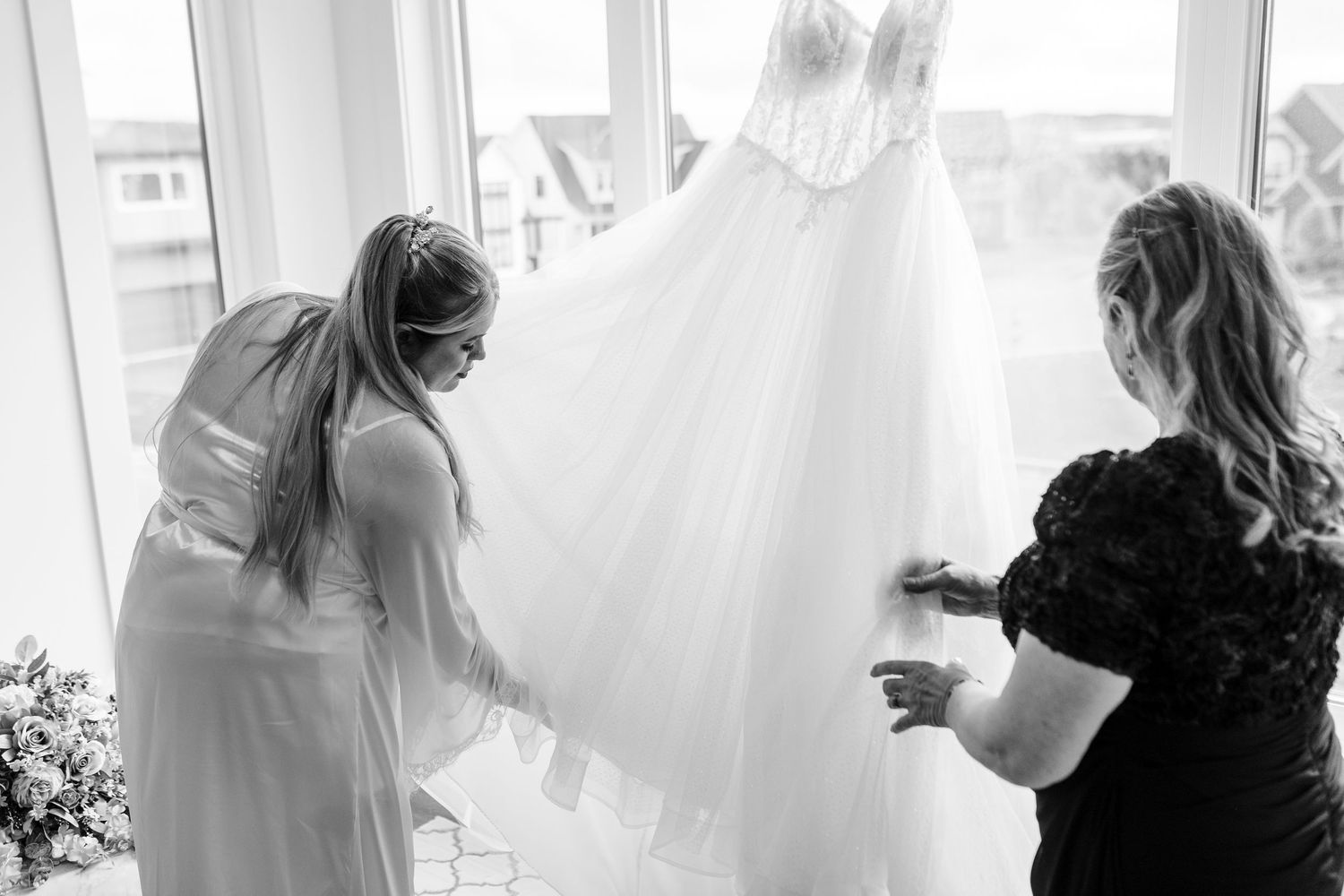 Two people carefully adjusting a flowing white wedding dress near a bright window in black and white.