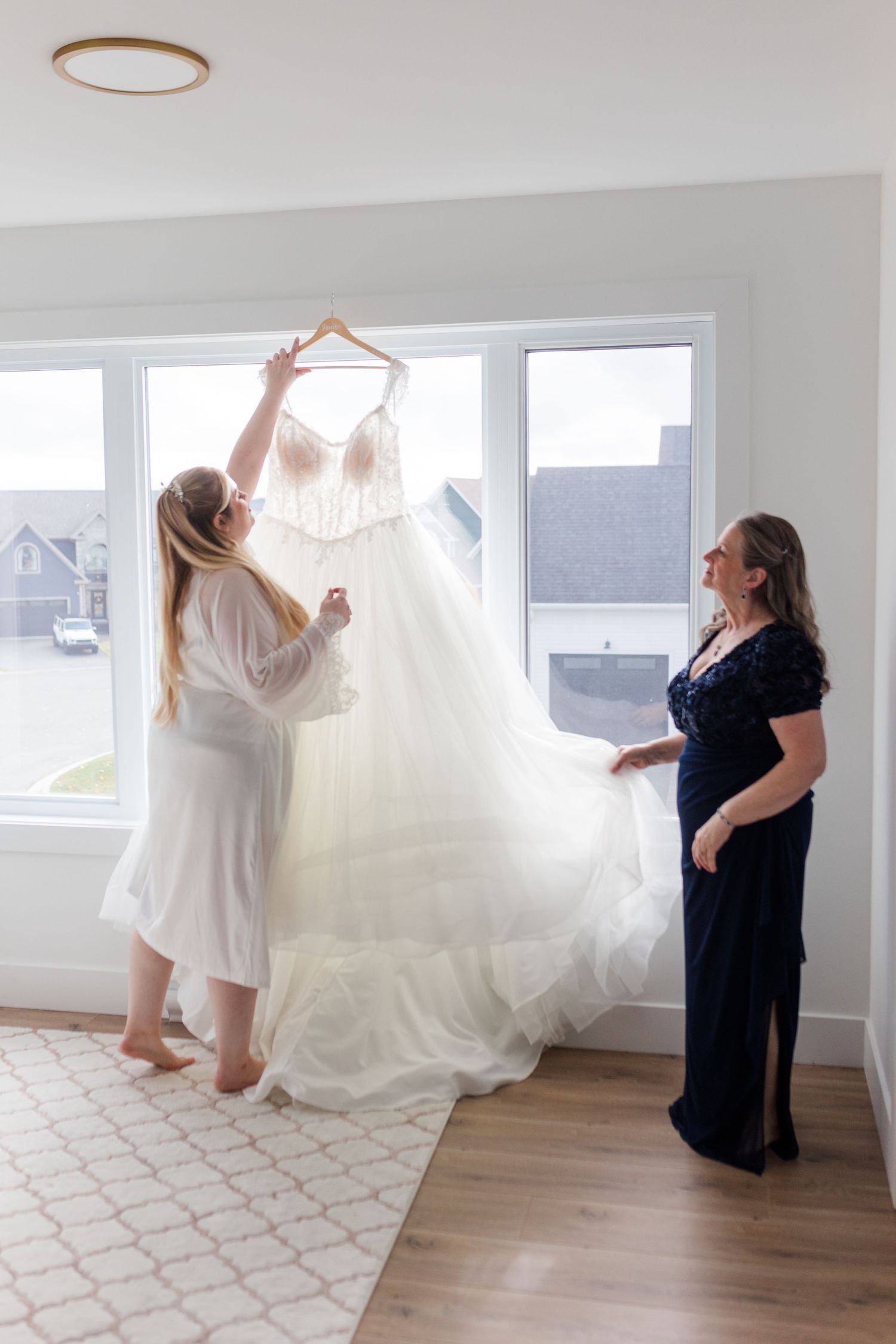 Two people admiring an elegant white wedding gown hanging in a bright room with large windows.