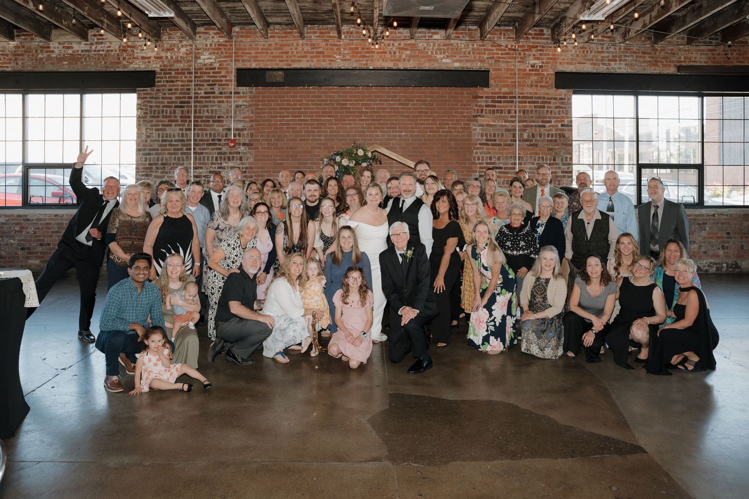 A panoramic sequence of wedding guests sitting on the floor in an industrial brick loft venue with large windows.