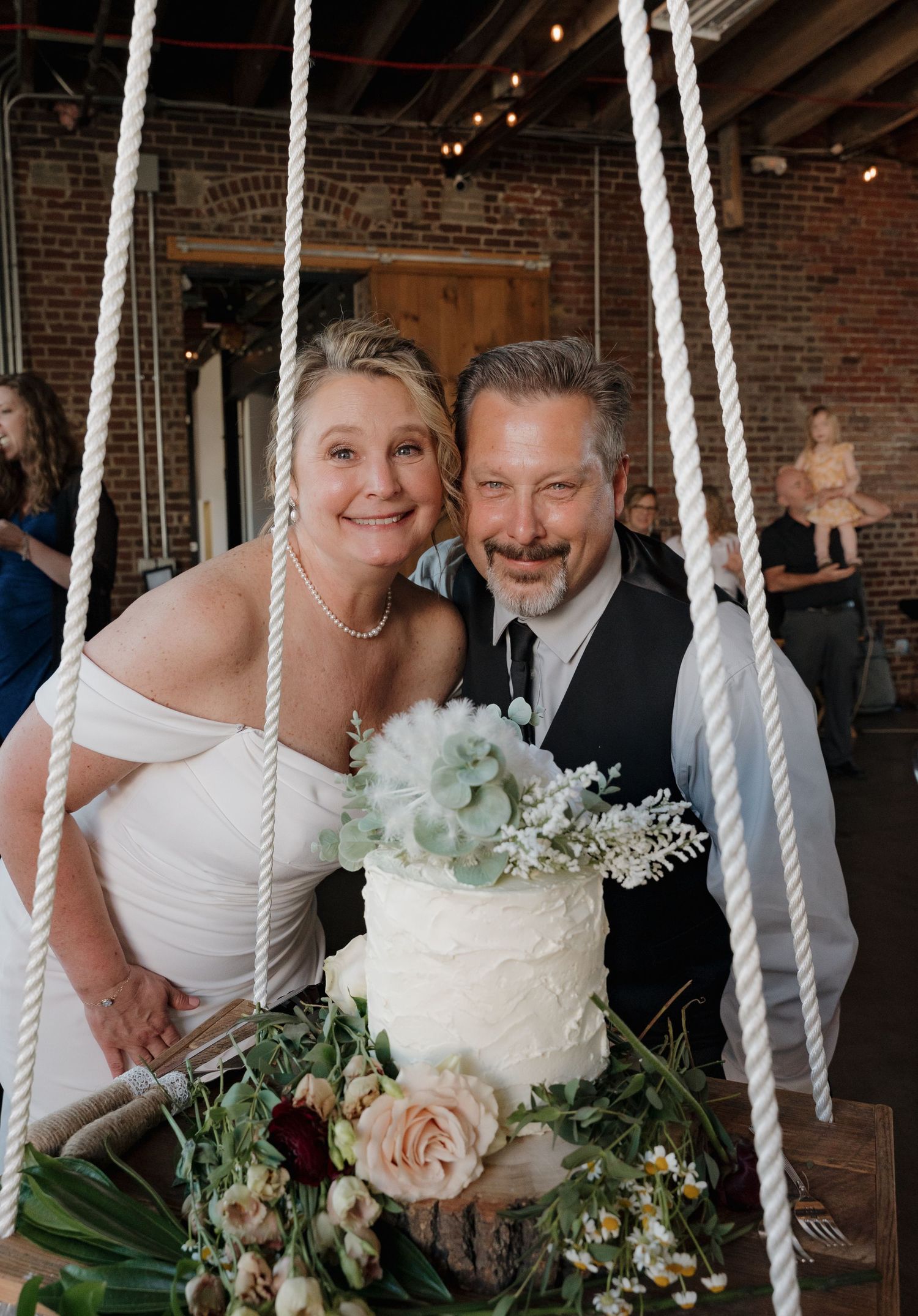 A rustic wedding cake hangs from white ropes in an industrial brick venue with soft lighting and floral decorations.