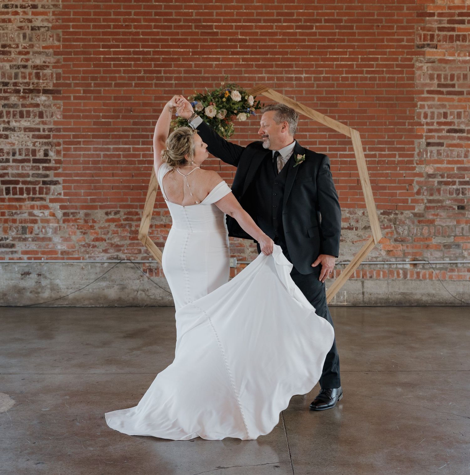 Couple performs elegant dance moves in front of rustic brick wall, bride's white dress flowing gracefully during spin.