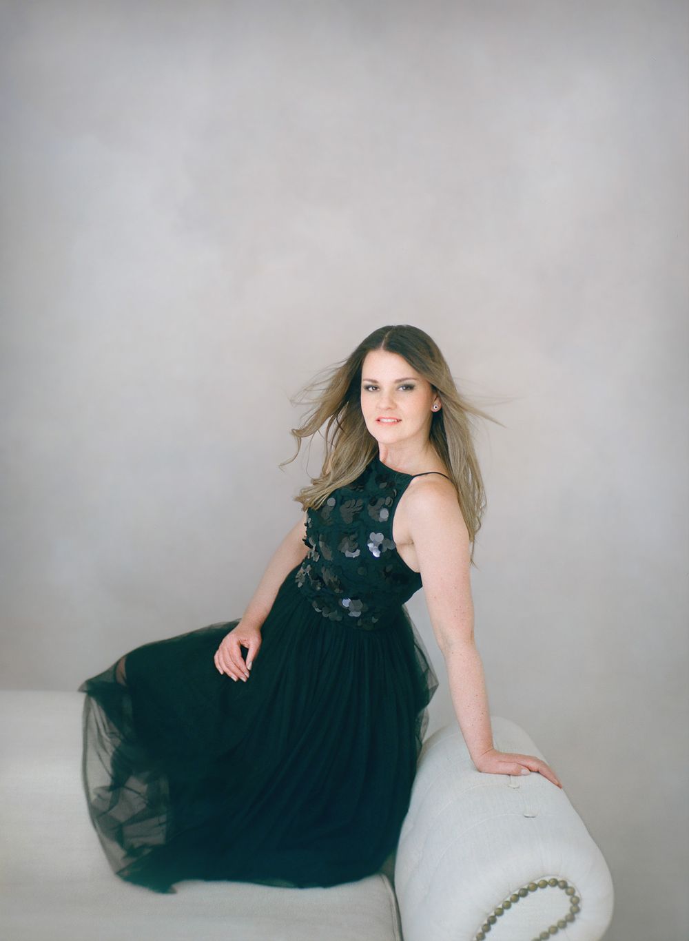 Elegant pose in black formal dress resting against white furniture.