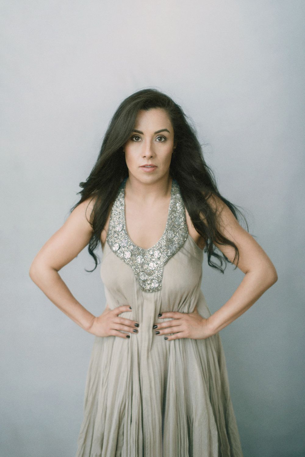 A fashion portrait with long dark hair in a flowing taupe dress with ornate silver embellished neckline against grey background.