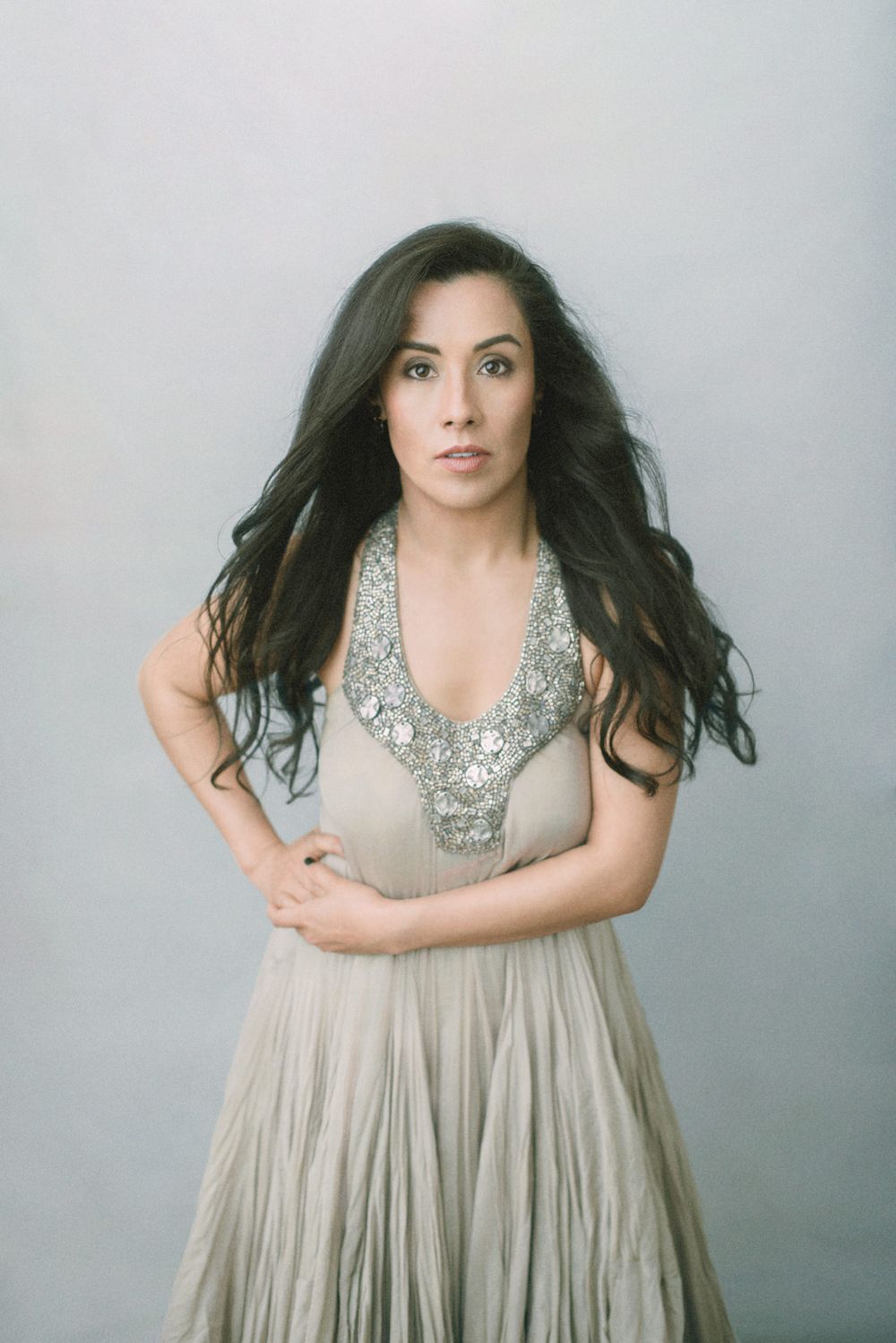 Studio portrait of someone in flowy beige dress with sequined neckline and long dark hair against light background.
