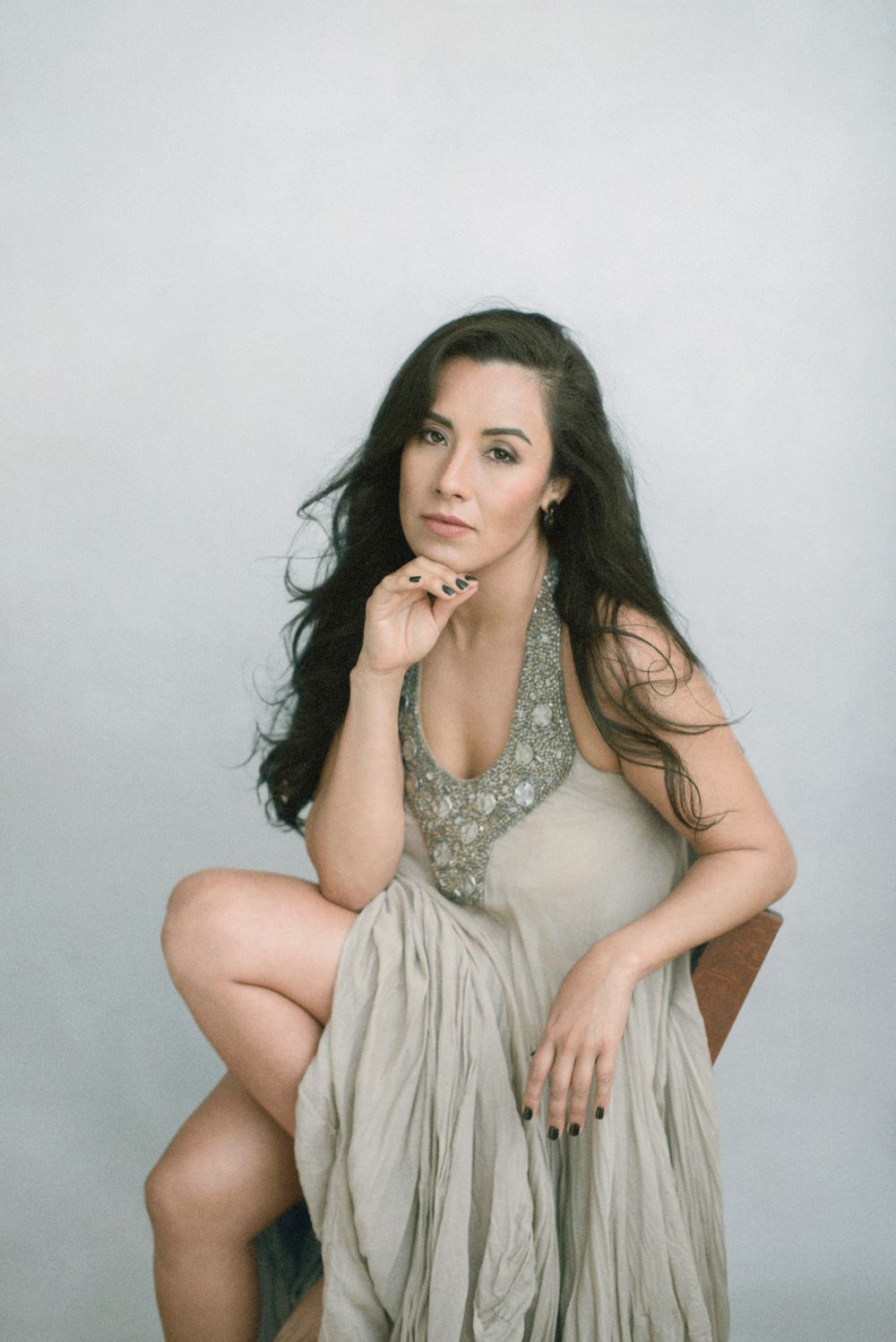 A model in a flowing beige dress with embellished neckline poses thoughtfully on a simple grey background.