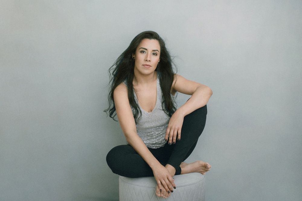 Model sits casually in a gray tank top and dark pants against a light gray studio backdrop.