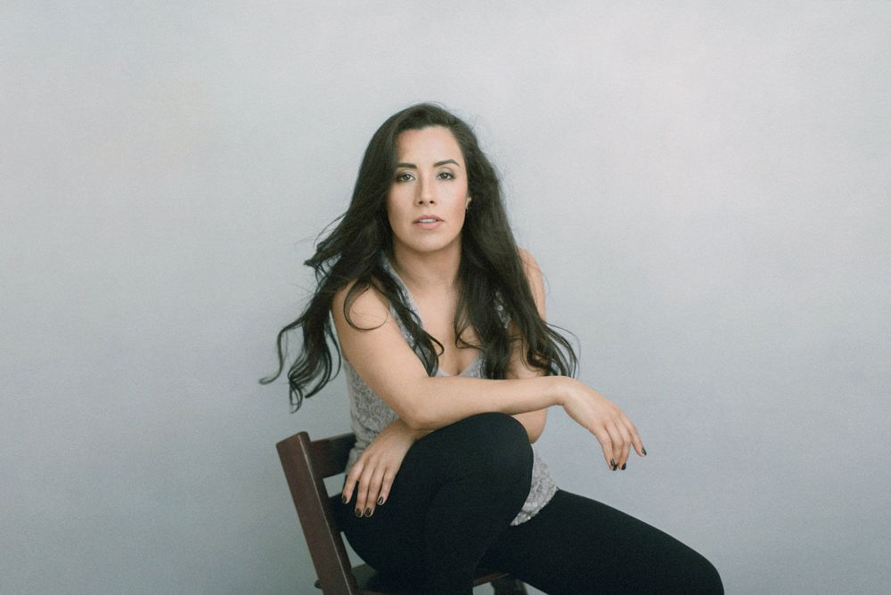 Portrait of dark-haired woman in casual attire sitting on wooden chair against neutral backdrop.