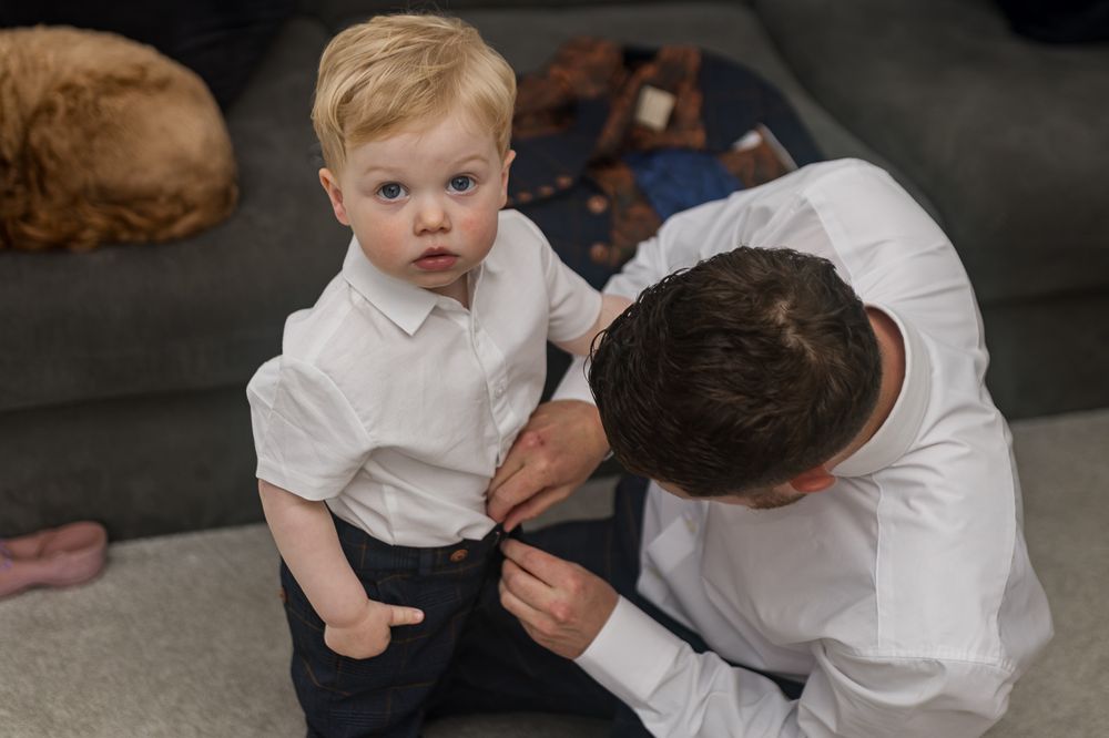 A toddler in formal white attire interacts with someone in a navy suit.