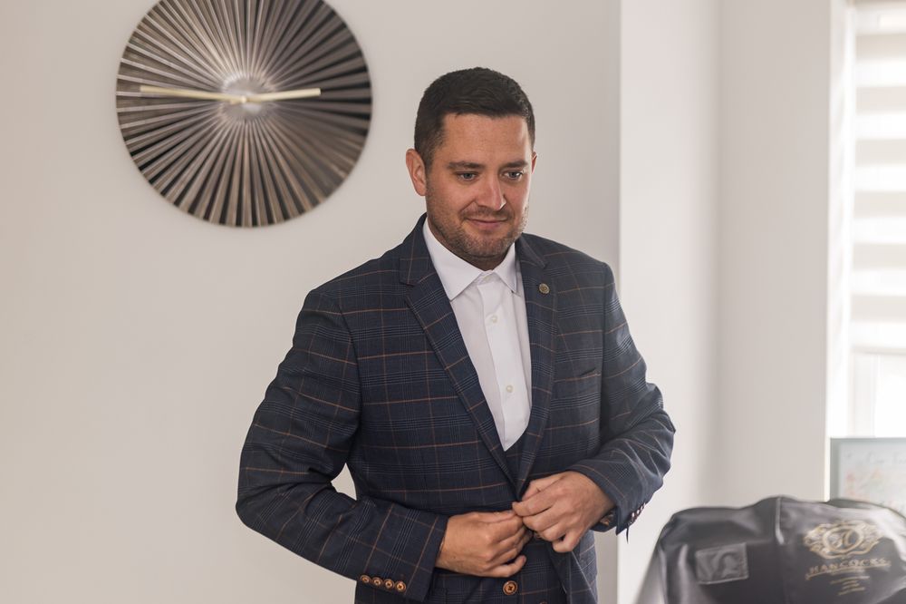 A person adjusts their navy checkered suit jacket while standing in a bright room with modern decor.