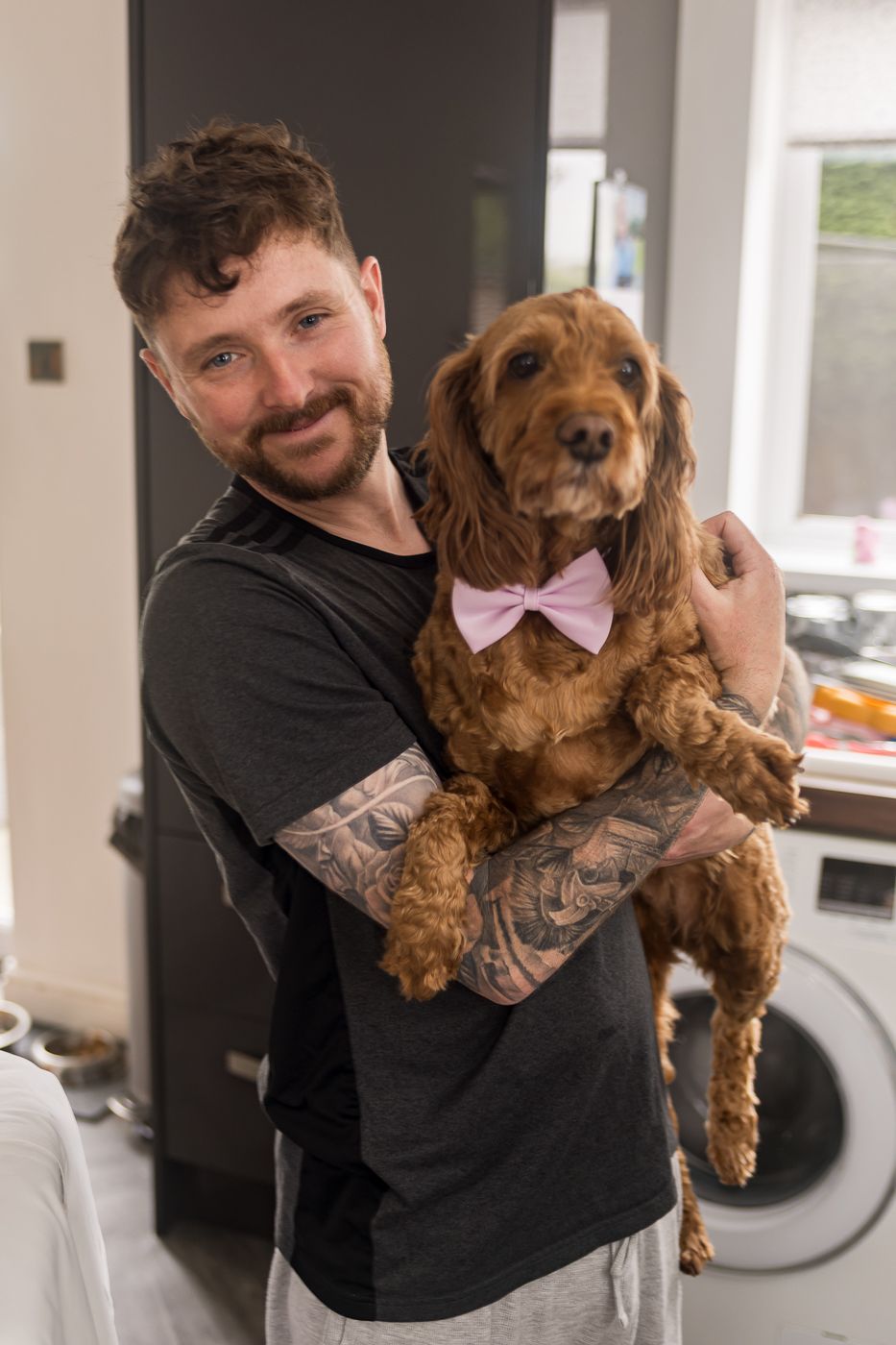 Person in black shirt holding a brown cockapoo dog wearing a pink bowtie.