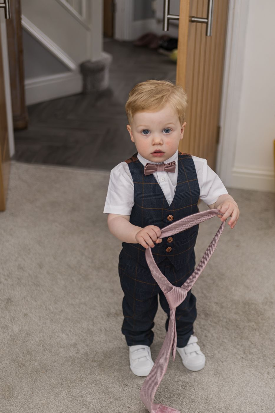 A small child in a navy vest and white shirt plays with a pink tie on a gray carpet.