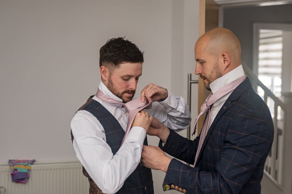 Two people in formal suits help each other with their ties in a bright room.