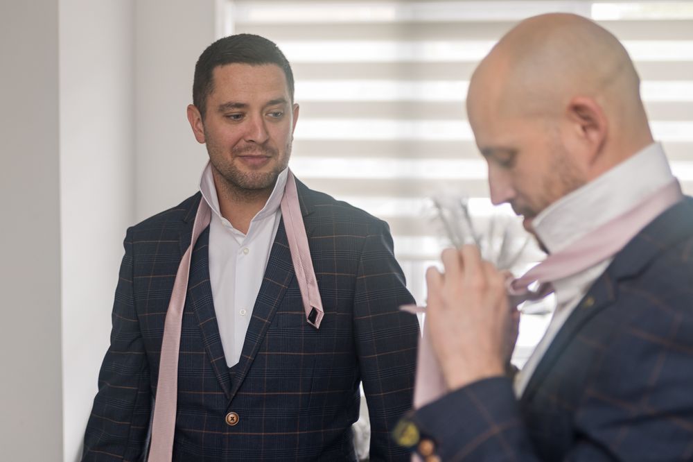 Two people in dark suits assist each other with formal attire preparations.