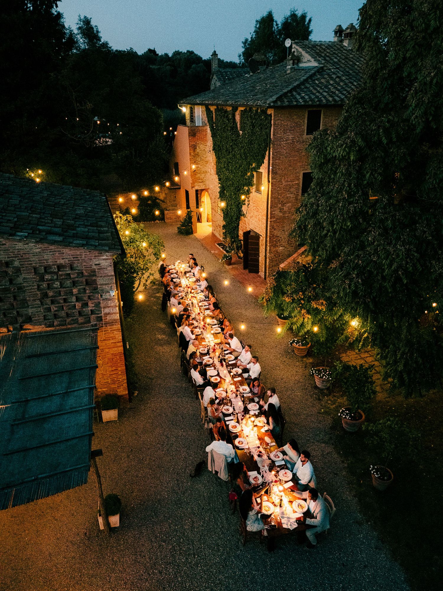 Aerial view of a long outdoor dinner party table illuminated by string lights at dusk in a rustic courtyard setting.