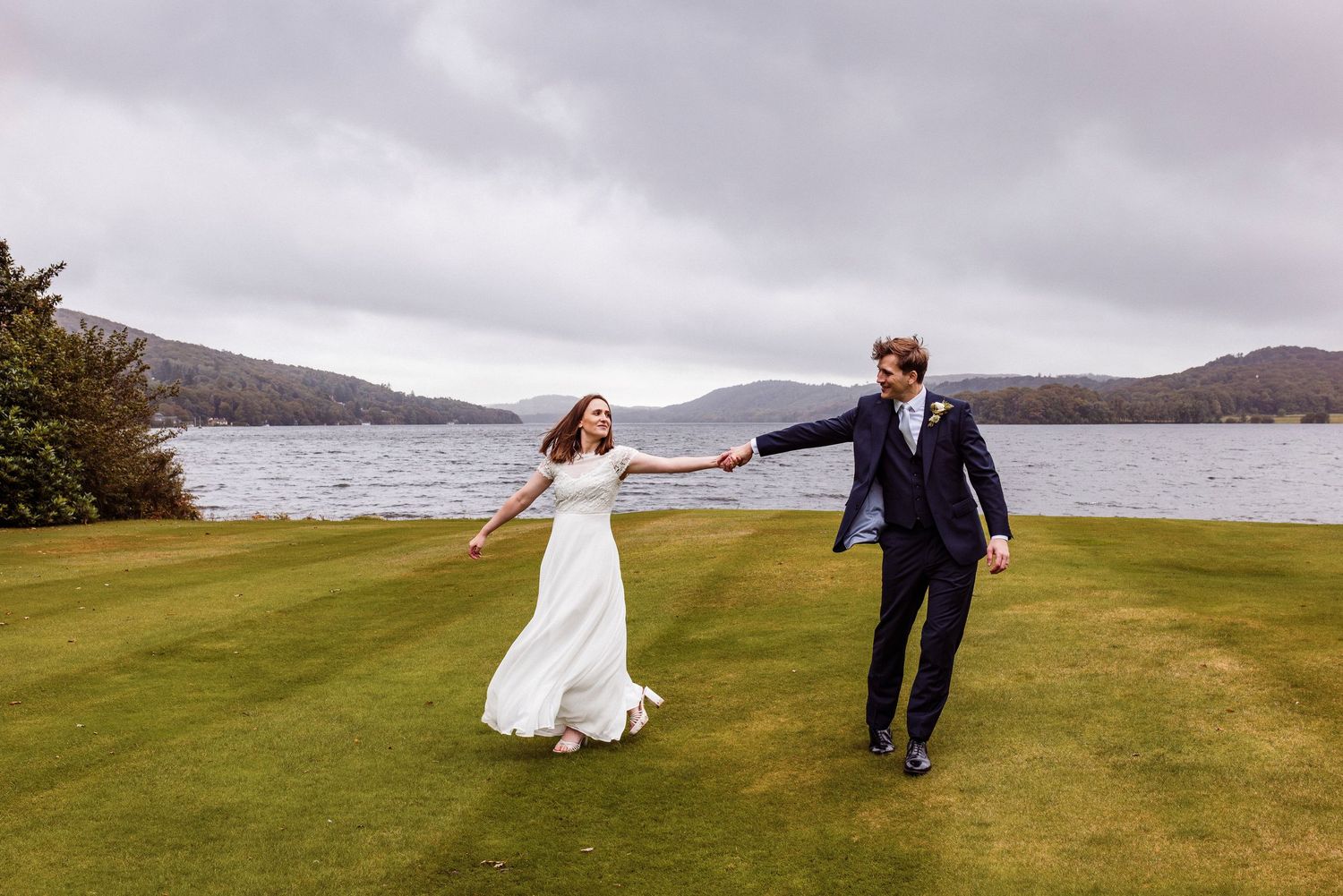 Couple in wedding attire playfully running and dancing across a scenic green hillside overlooking a misty coastline.