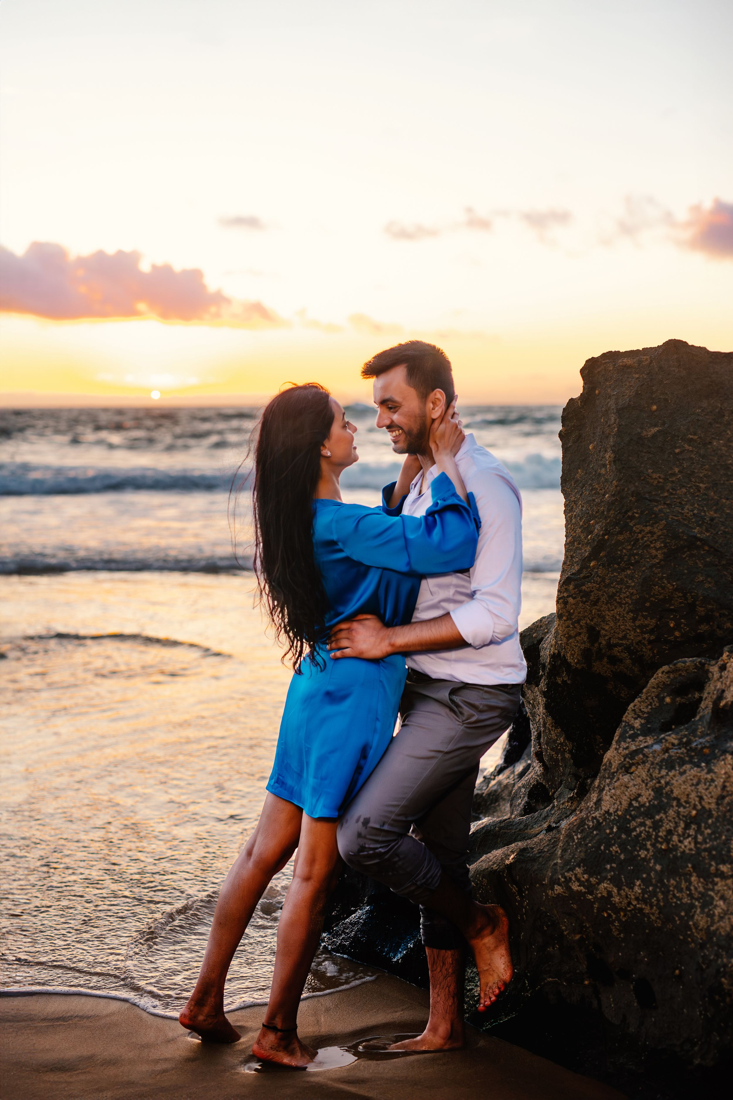 A romantic couple sits together on rocky beach during a vibrant sunset.