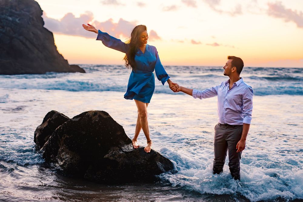 Two people share a romantic moment on coastal rocks at sunset as ocean waves crash around them.