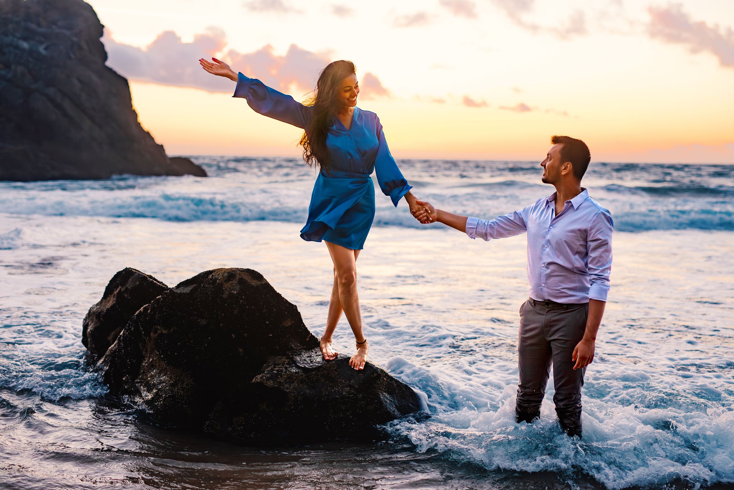 Two people share a romantic moment on coastal rocks at sunset as ocean waves crash around them.