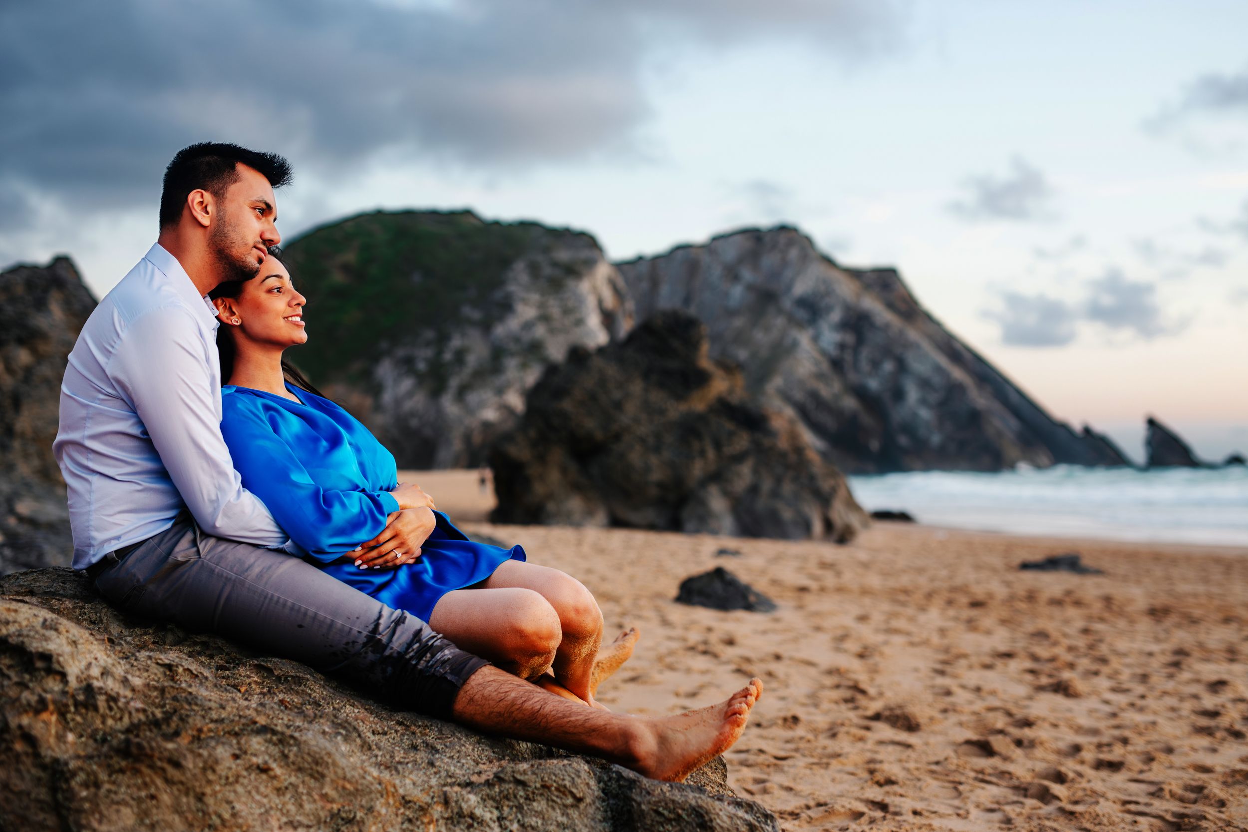 Couple sitting together on rocky beach cliffs during sunset overlooking the ocean waves.