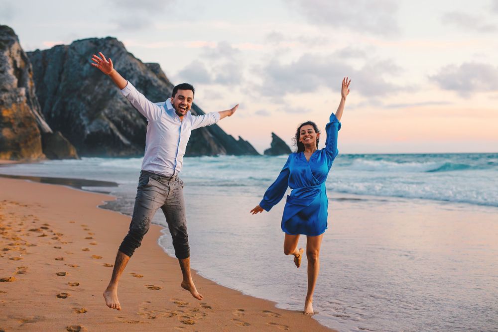 People enjoy a playful moment together on a sandy beach at sunset with waves and cliffs in the background.